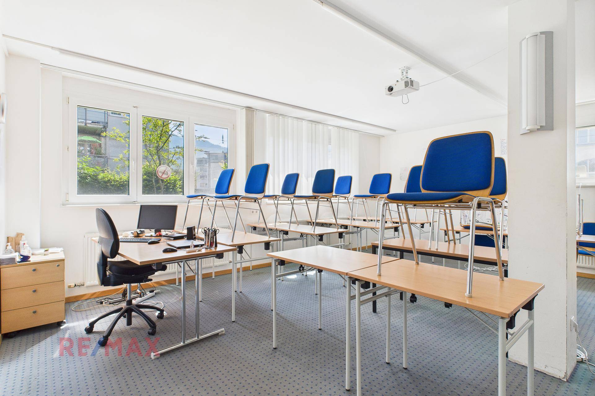 Klassenzimmer mit aufgestellten Tischen und Stühlen, einem Schreibtisch und großen Fenstern.