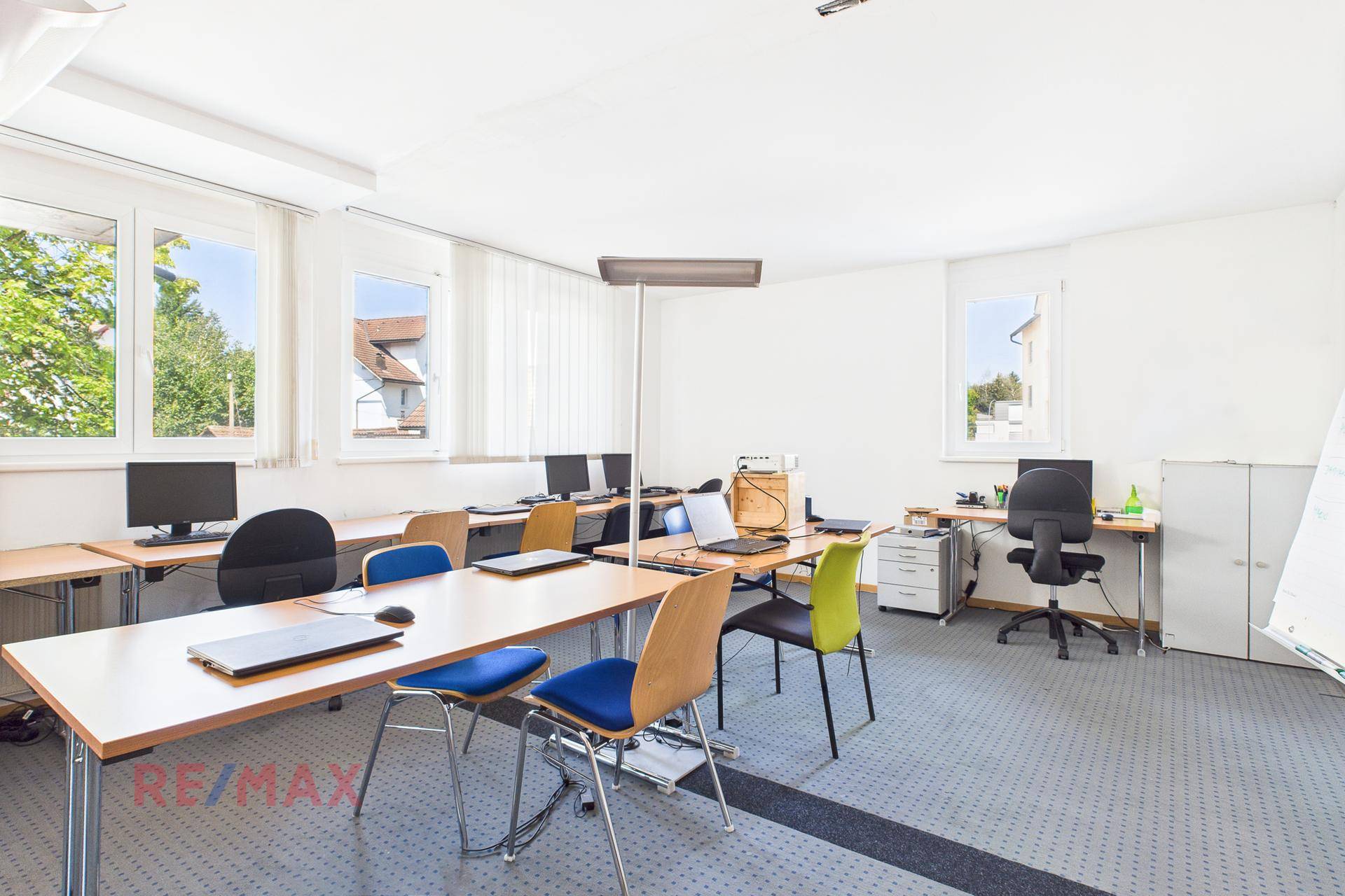 Büroraum mit mehreren Arbeitsplätzen, Computern und großen Fenstern, die viel Licht hereinlassen.