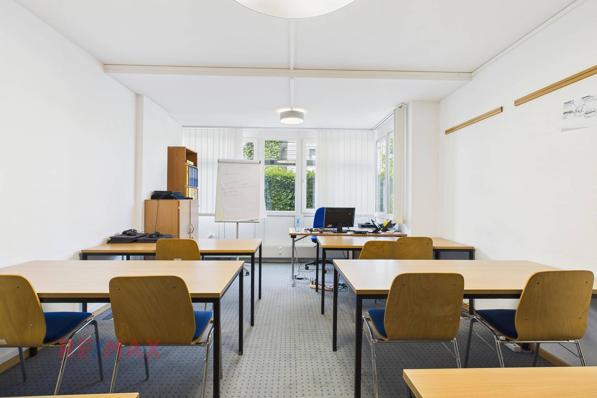 Klassenzimmer mit mehreren Tischen und Stühlen, einer weißen Tafel und großen Fenstern.