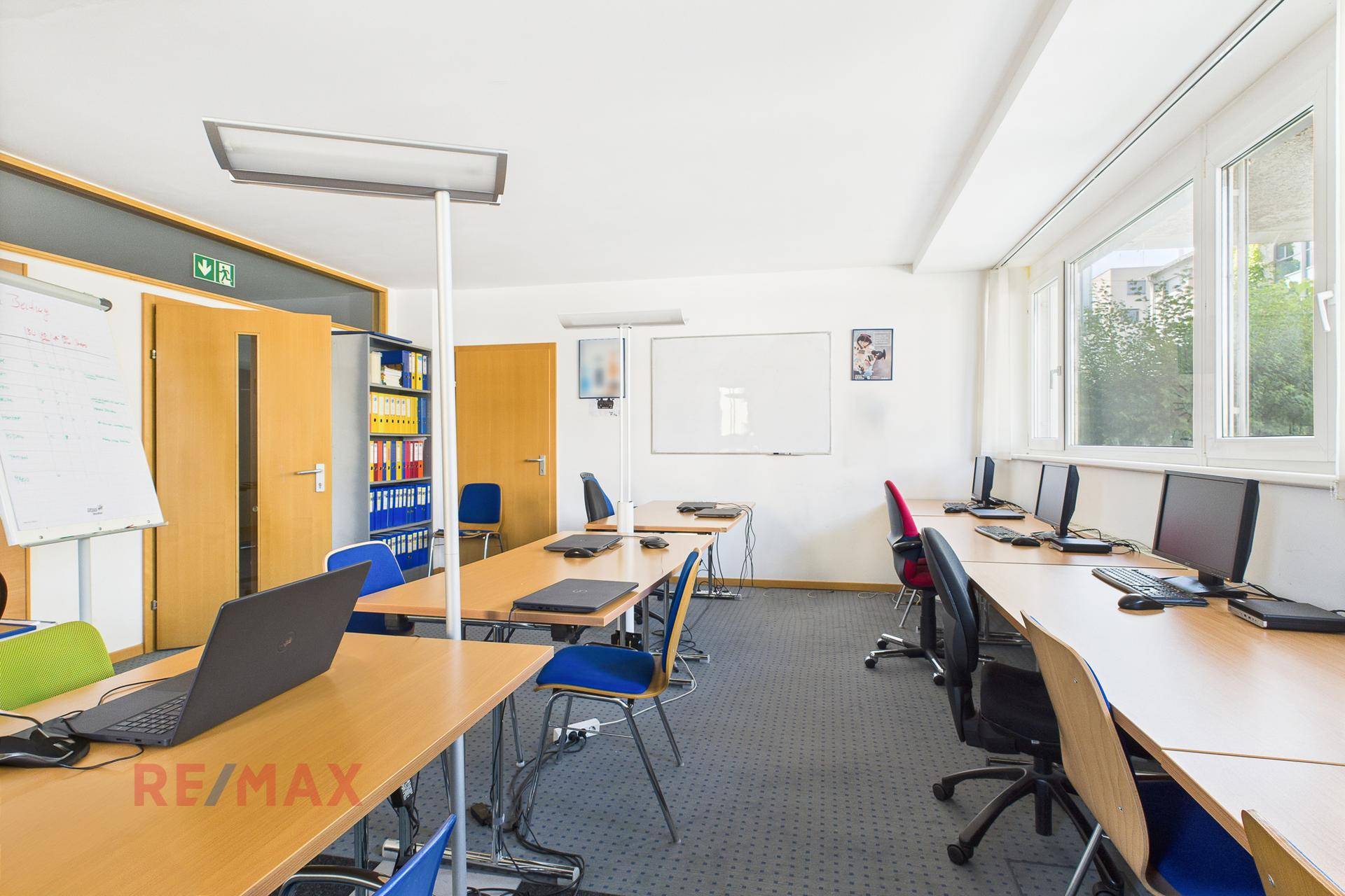 Büroraum mit mehreren Arbeitsplätzen, Computern und großen Fenstern, die viel Licht hereinlassen.