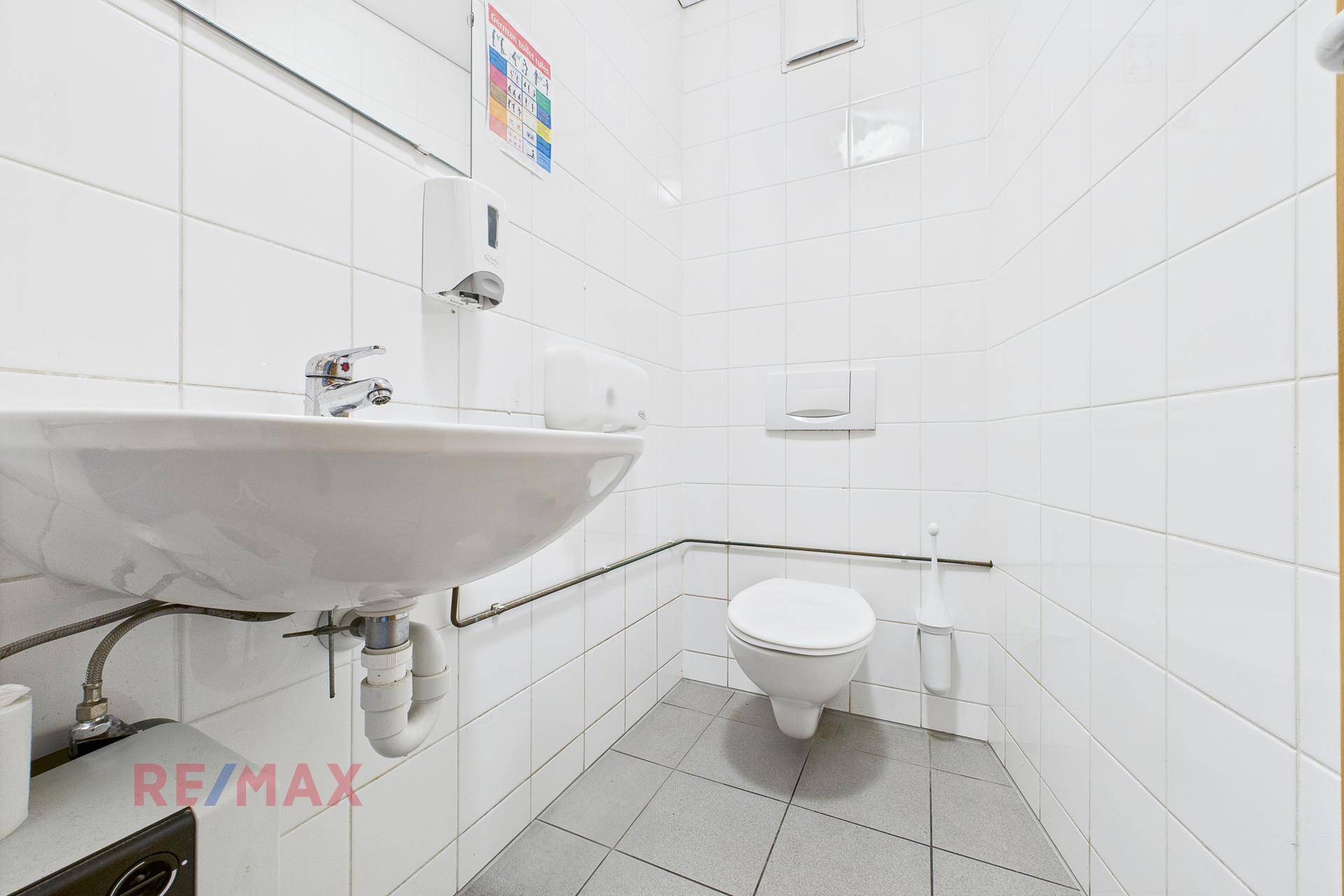 Saubere Toilette mit weißen Fliesen, Waschbecken und Wand-WC, ausgestattet mit Haltegriffen.