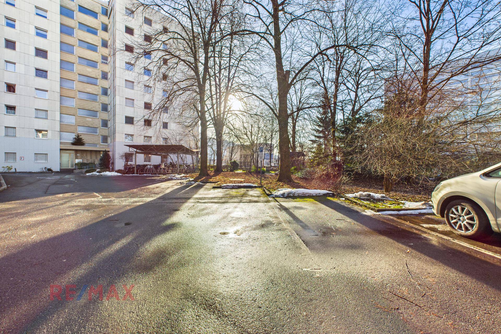 Außenbereich mit Parkplätzen und Bäumen vor einem mehrstöckigen Gebäude.