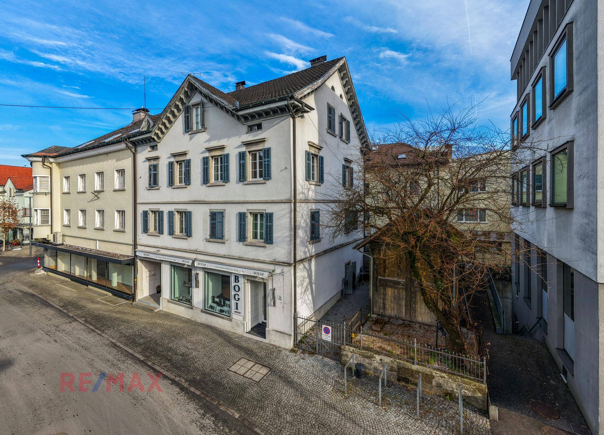 Ansicht des historischen Gebäudes mit Kopfsteinpflasterstraße und umliegender Bebauung unter blauem Himmel.