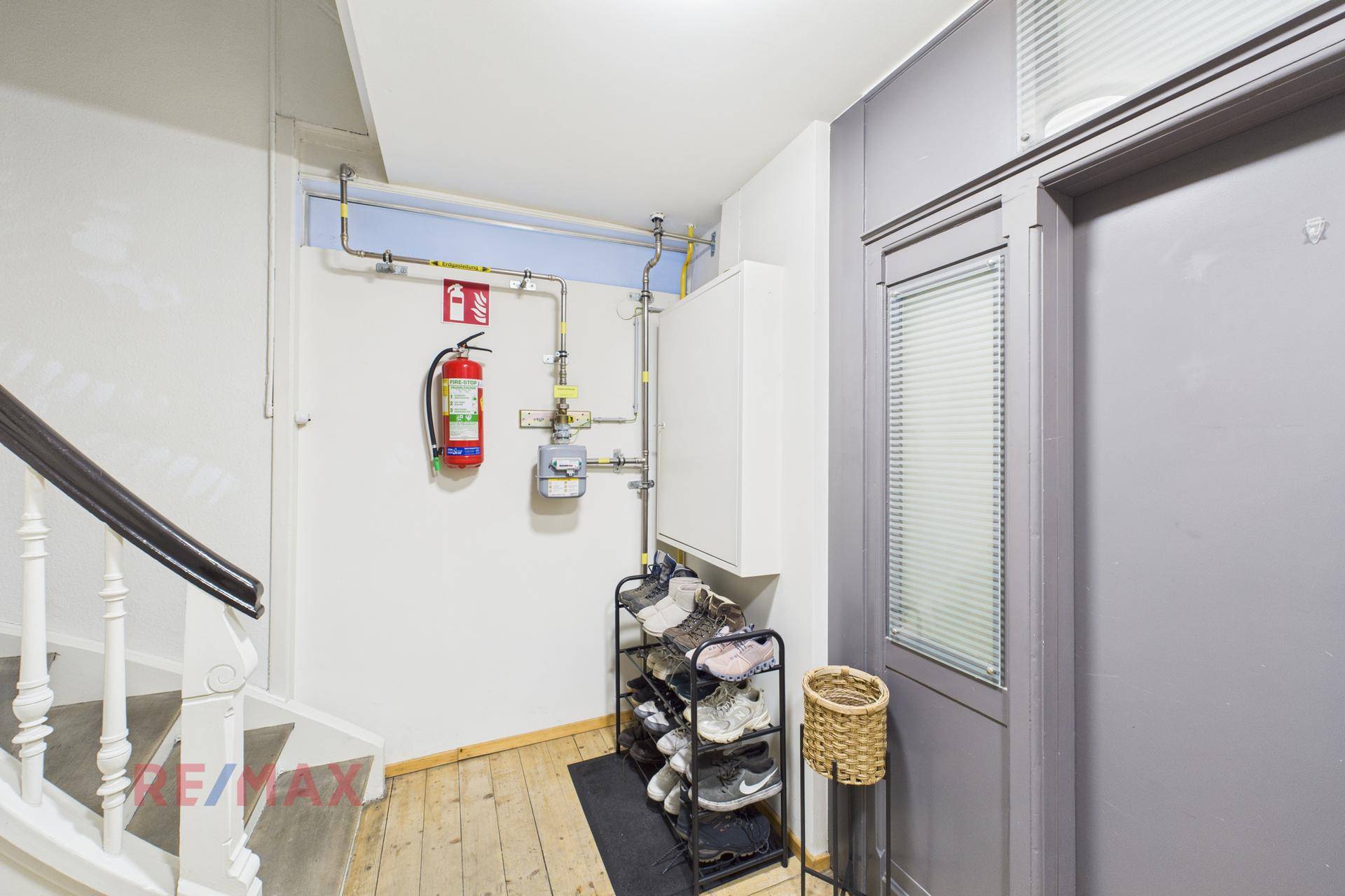 Treppenbereich mit sichtbaren Rohren, Feuerlöscher und Schuhregal, typisch für einen Altbau.