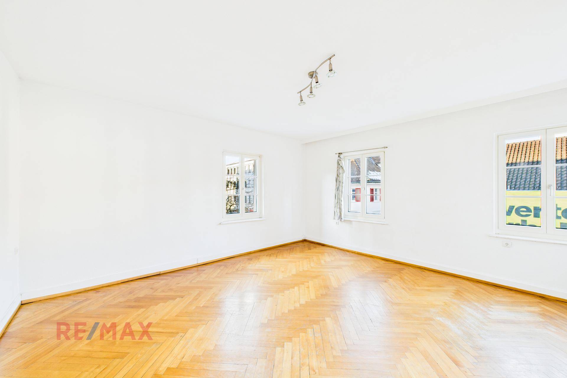 Geräumiges Wohnzimmer mit Fischgrätparkett und weißen Wänden, mit Blick nach draußen.
