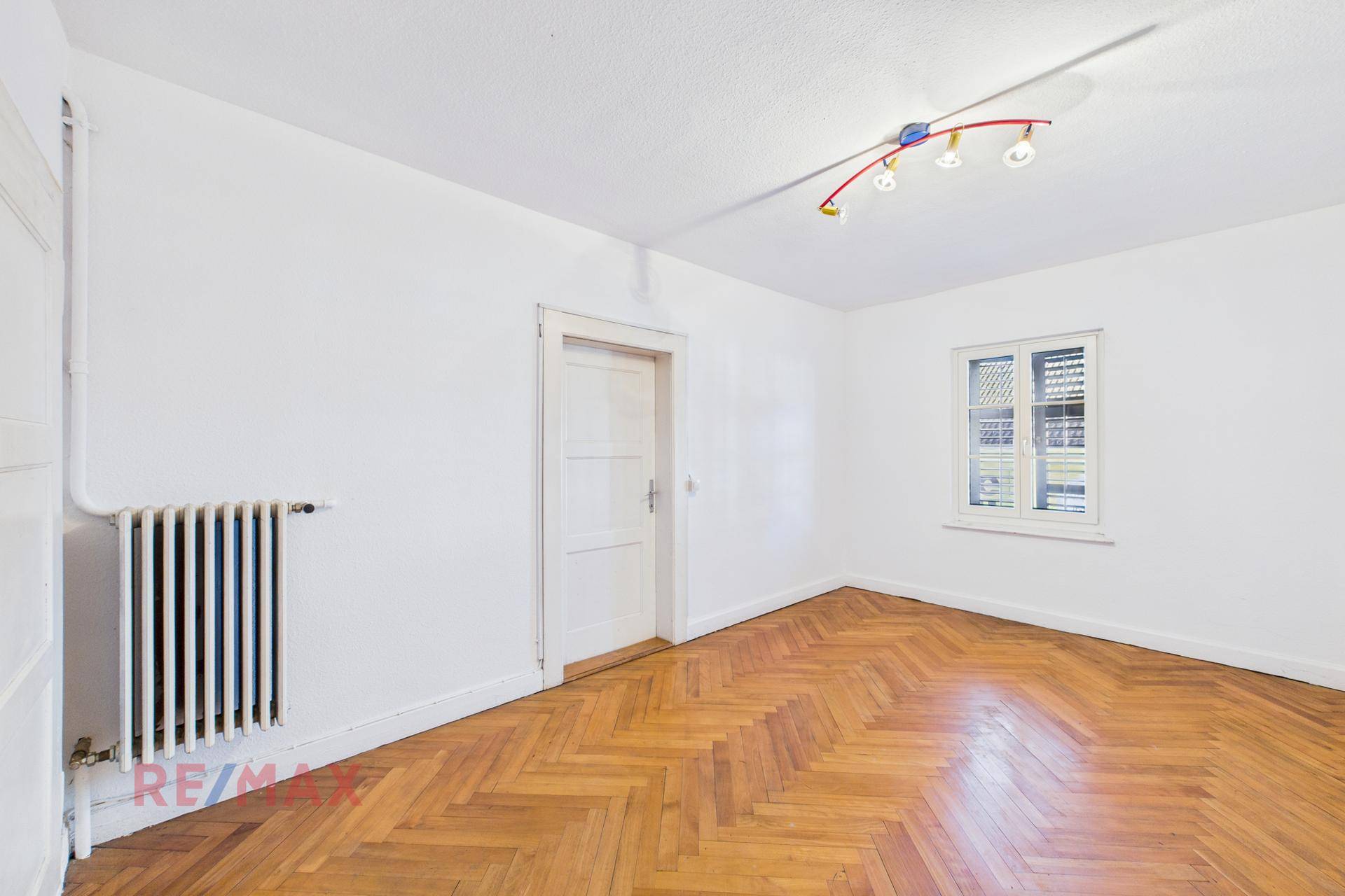 Geräumiges Wohnzimmer mit Fischgrätparkett und weißen Wänden, mit Heizkörper.
