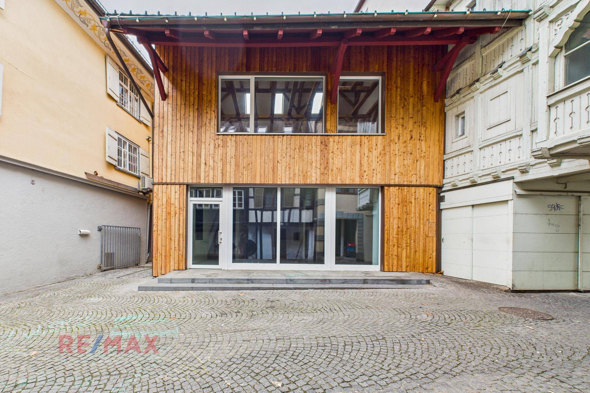 Moderne Holzfassade mit großen Schaufenstern und Eingangstür, umgeben von historischer Bebauung.