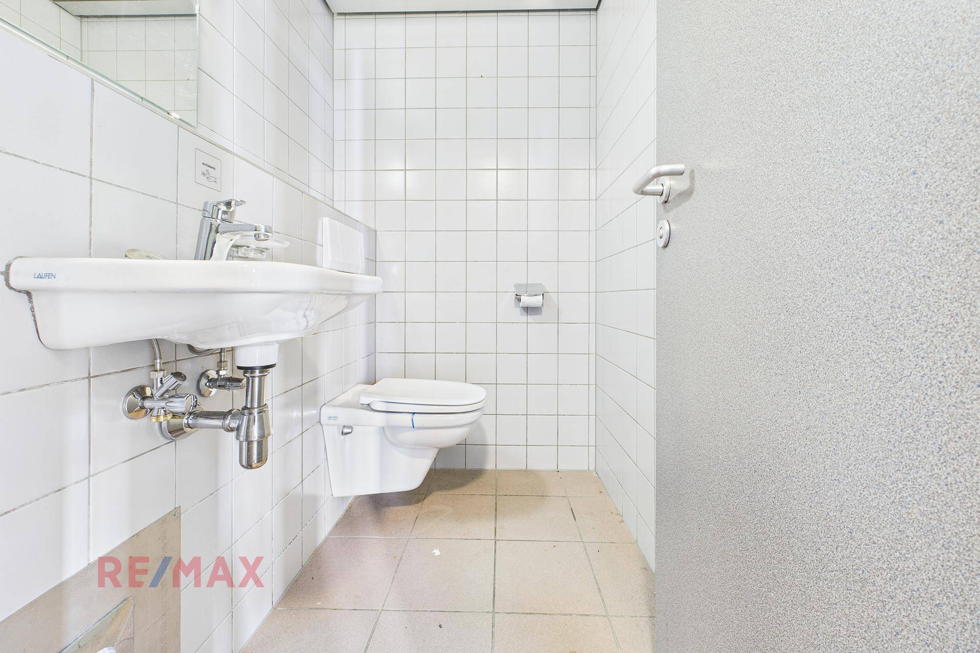 Einfache Toilette mit weißen Fliesen, Waschbecken und wandhängendem WC in funktionalem und sauberem Zustand.