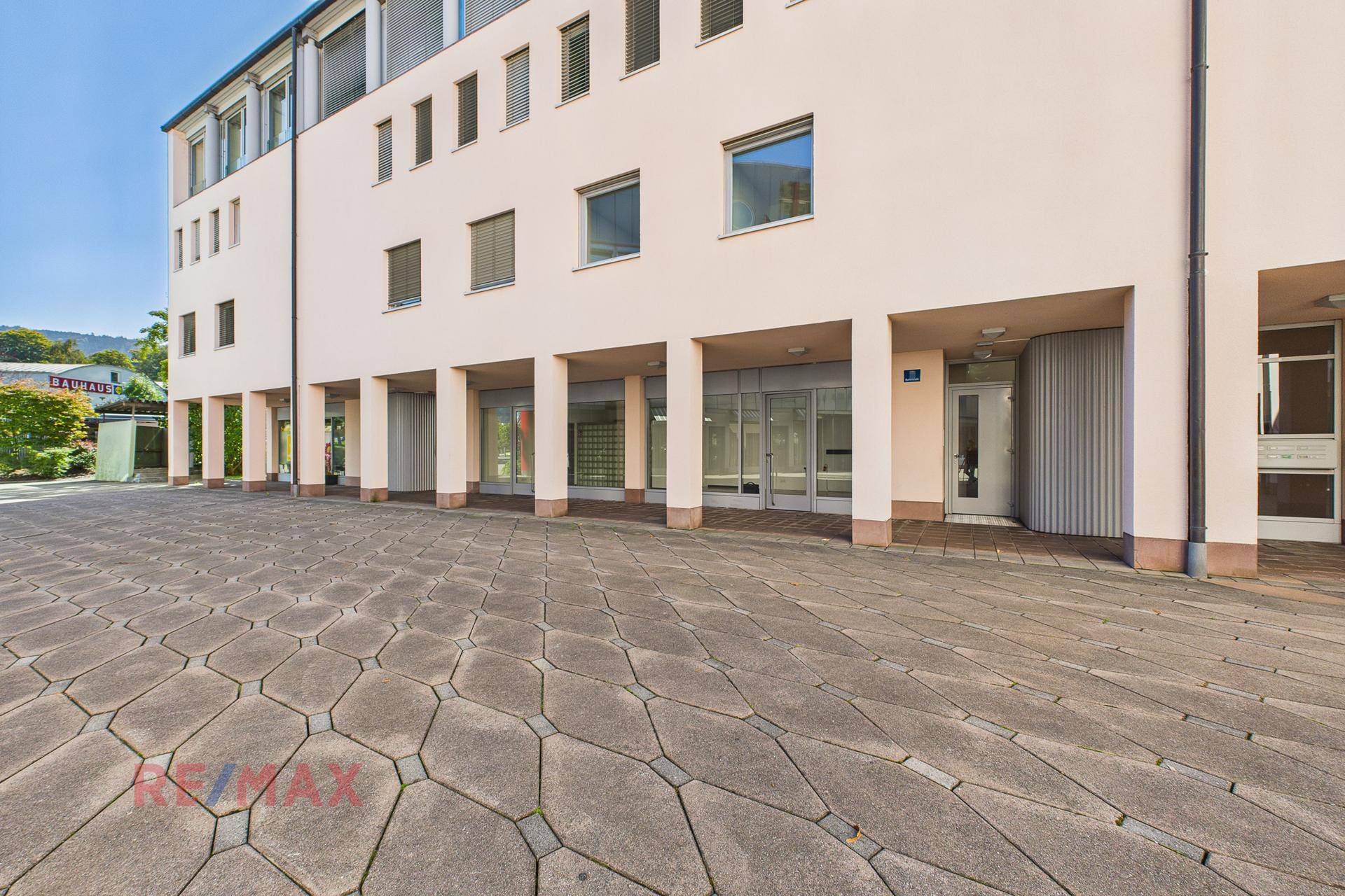 Gesamtansicht des modernen Gebäudes mit mehreren Etagen, großen Fenstern und einem weitläufigen gepflasterten Vorplatz.