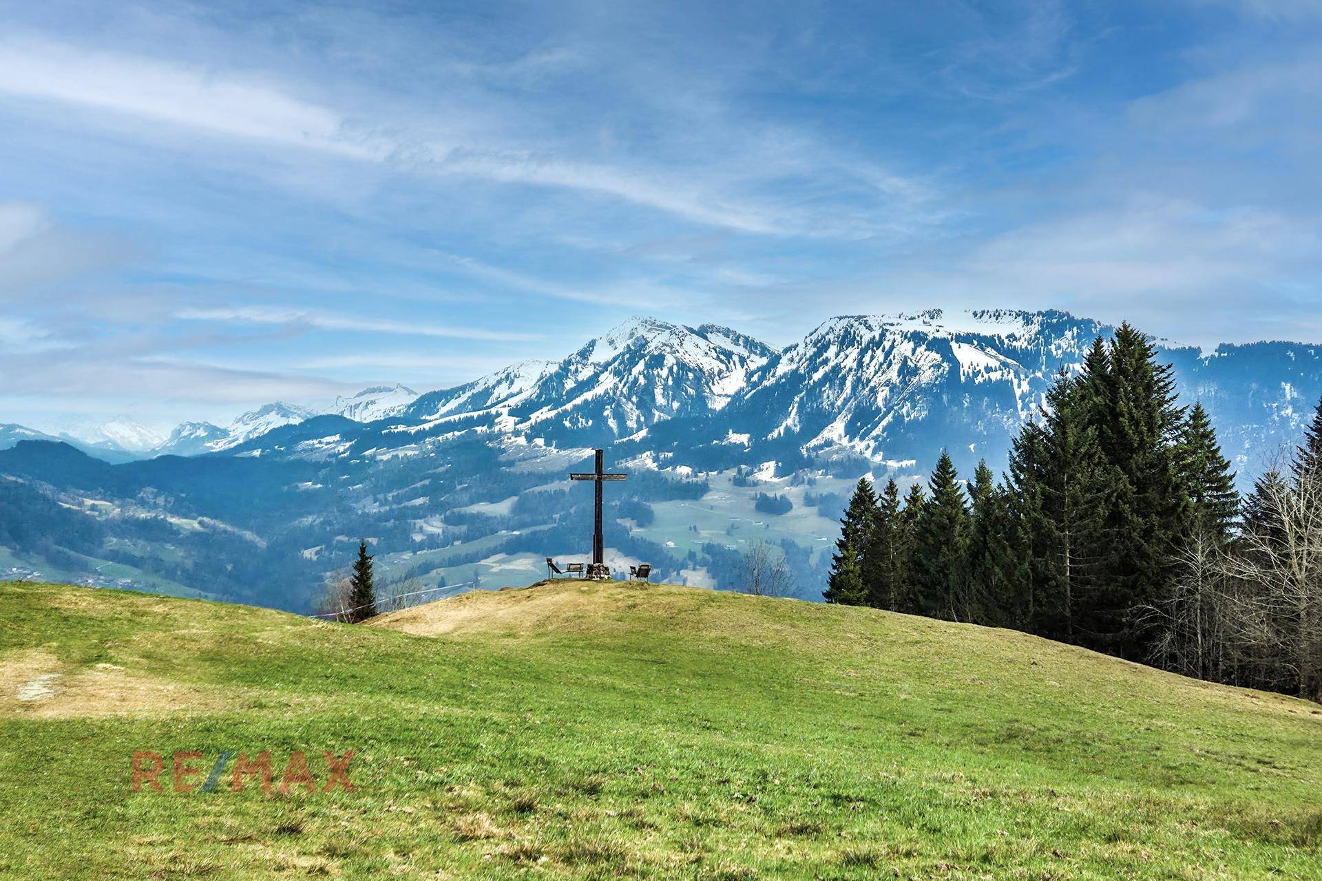 Weitläufige grüne Wiesen mit einem Gipfelkreuz und schneebedeckten Bergen im Hintergrund.