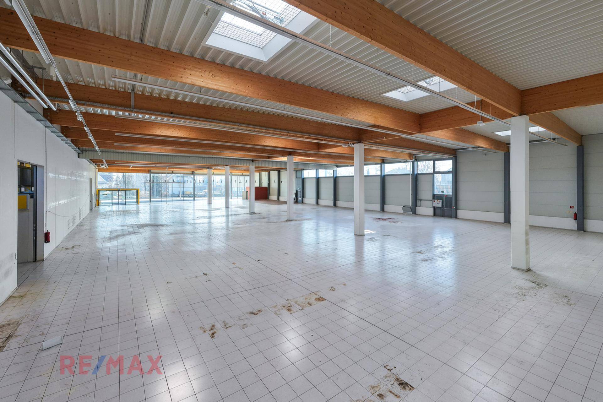 Geräumige, leere Halle mit Fliesenboden, hohen Decken, sichtbaren Holzträgern und großen Fenstern, ideal für Lager oder Produktion.