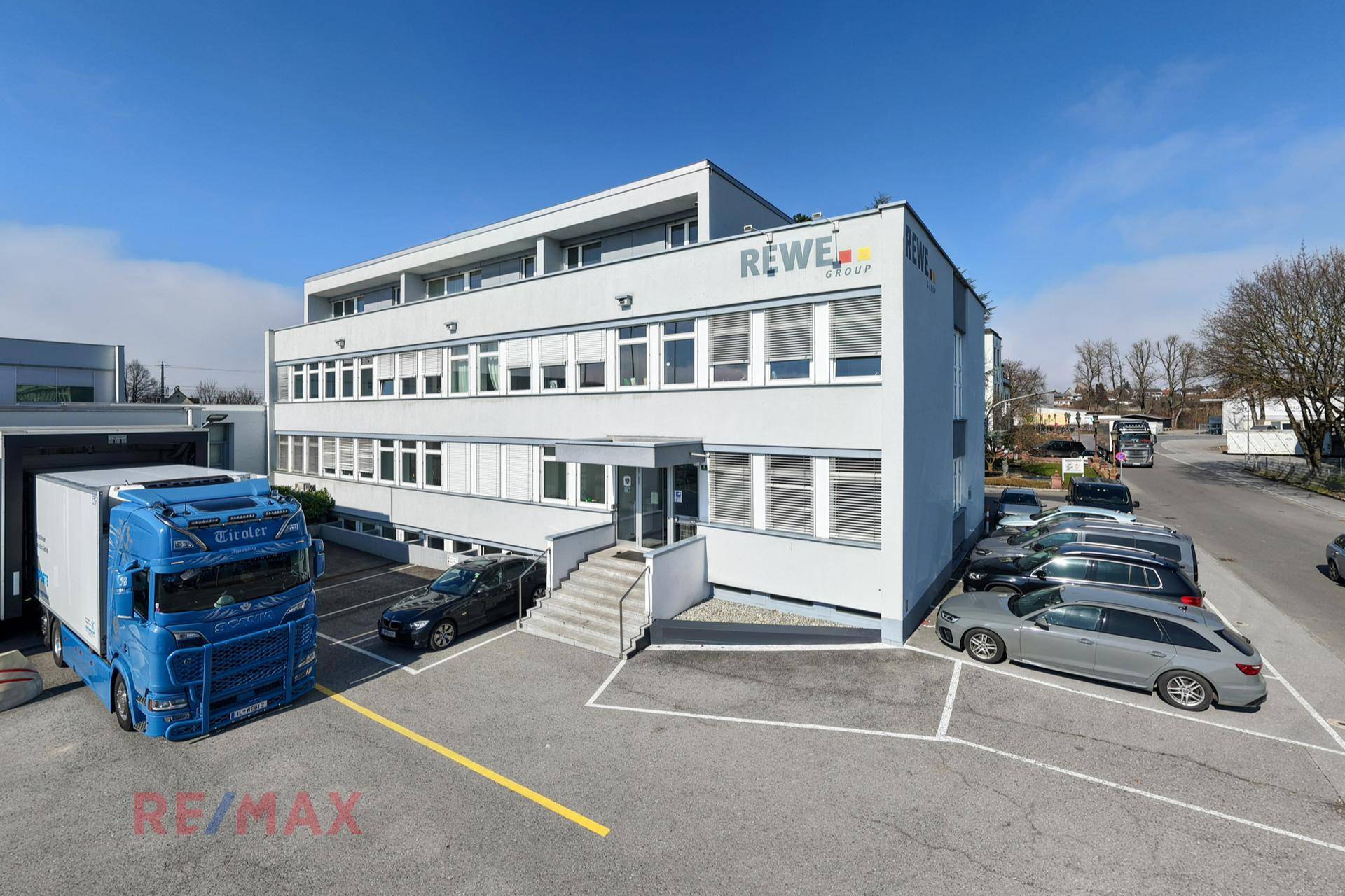 Modernes Bürogebäude mit mehreren Etagen, großen Fenstern und einem Parkplatz davor, unter blauem Himmel.