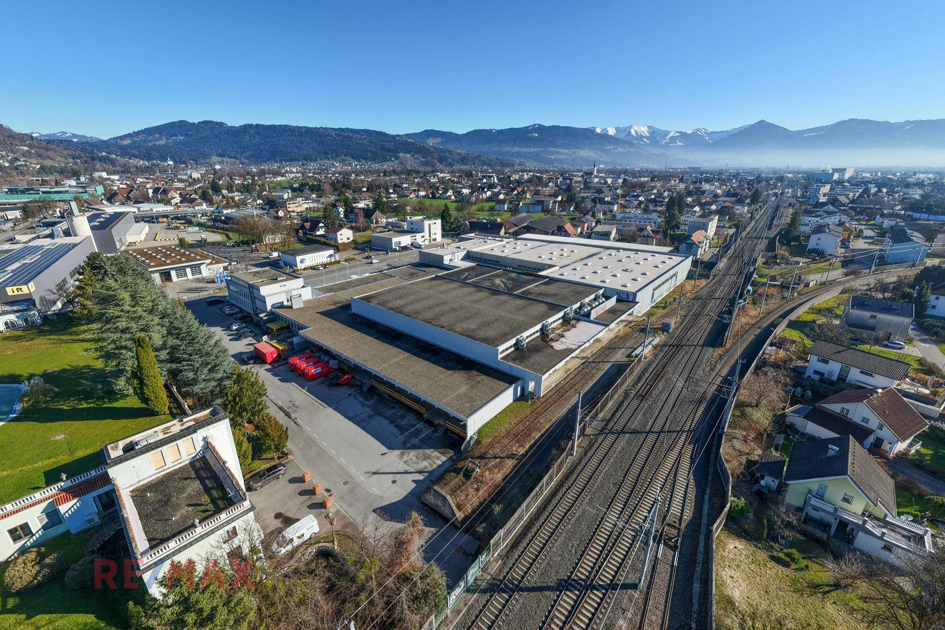 Luftaufnahme eines Gewerbegebiets mit großen Hallen, Bahngleisen und einer Bergkette im Hintergrund unter klarem Himmel.