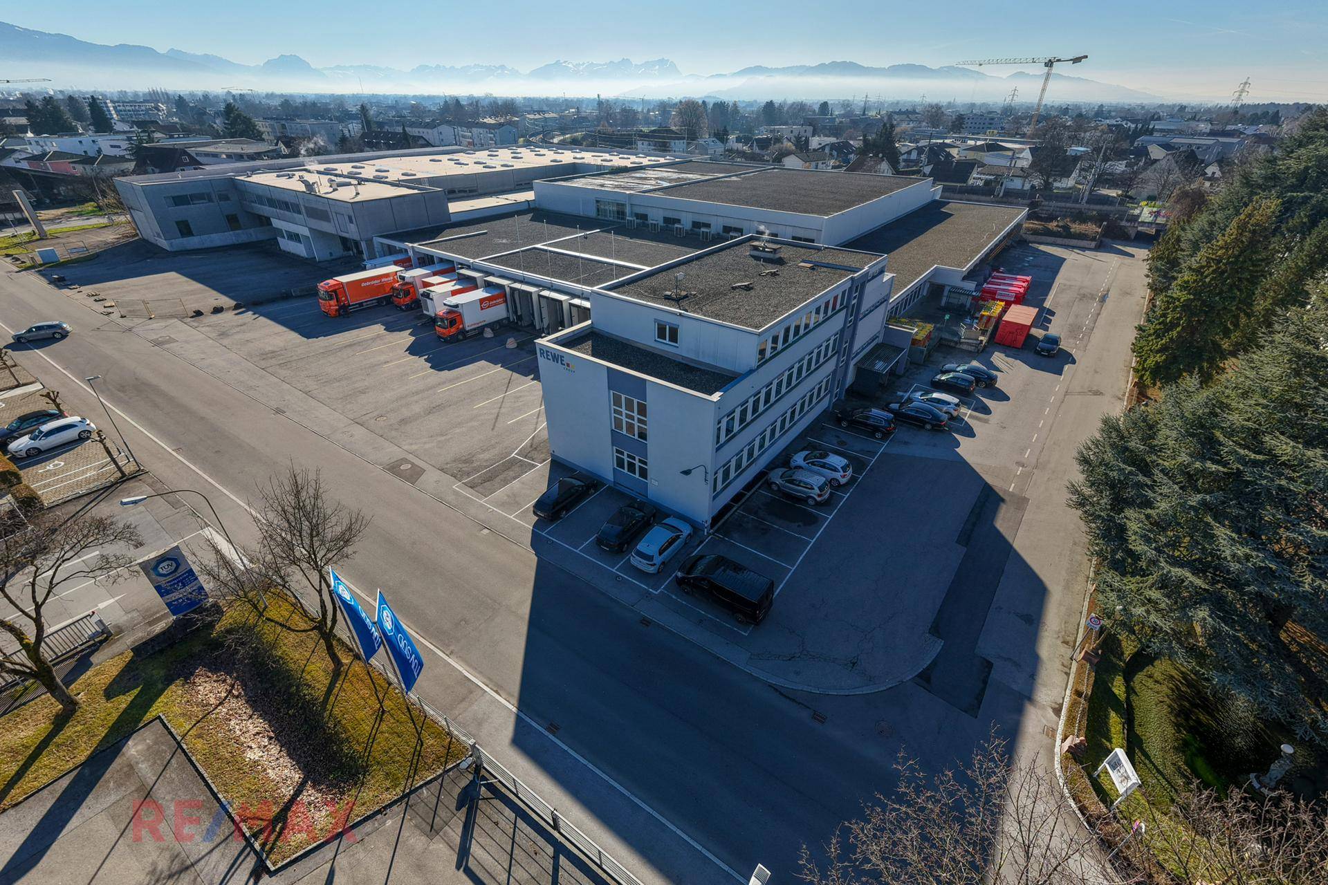 Übersicht aus der Luft eines großen Gewerbeareals mit Bürogebäude, Laderampen und Parkplätzen, mit Bergpanorama im Hintergrund.
