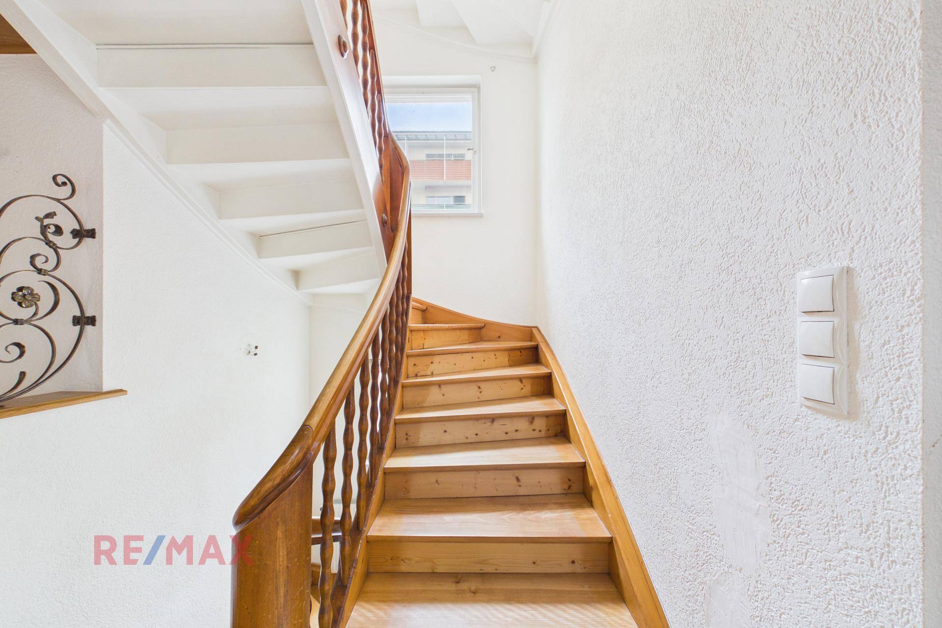 Holztreppe mit schmiedeeisernem Geländer in einem hellen Treppenhaus mit Fenster.