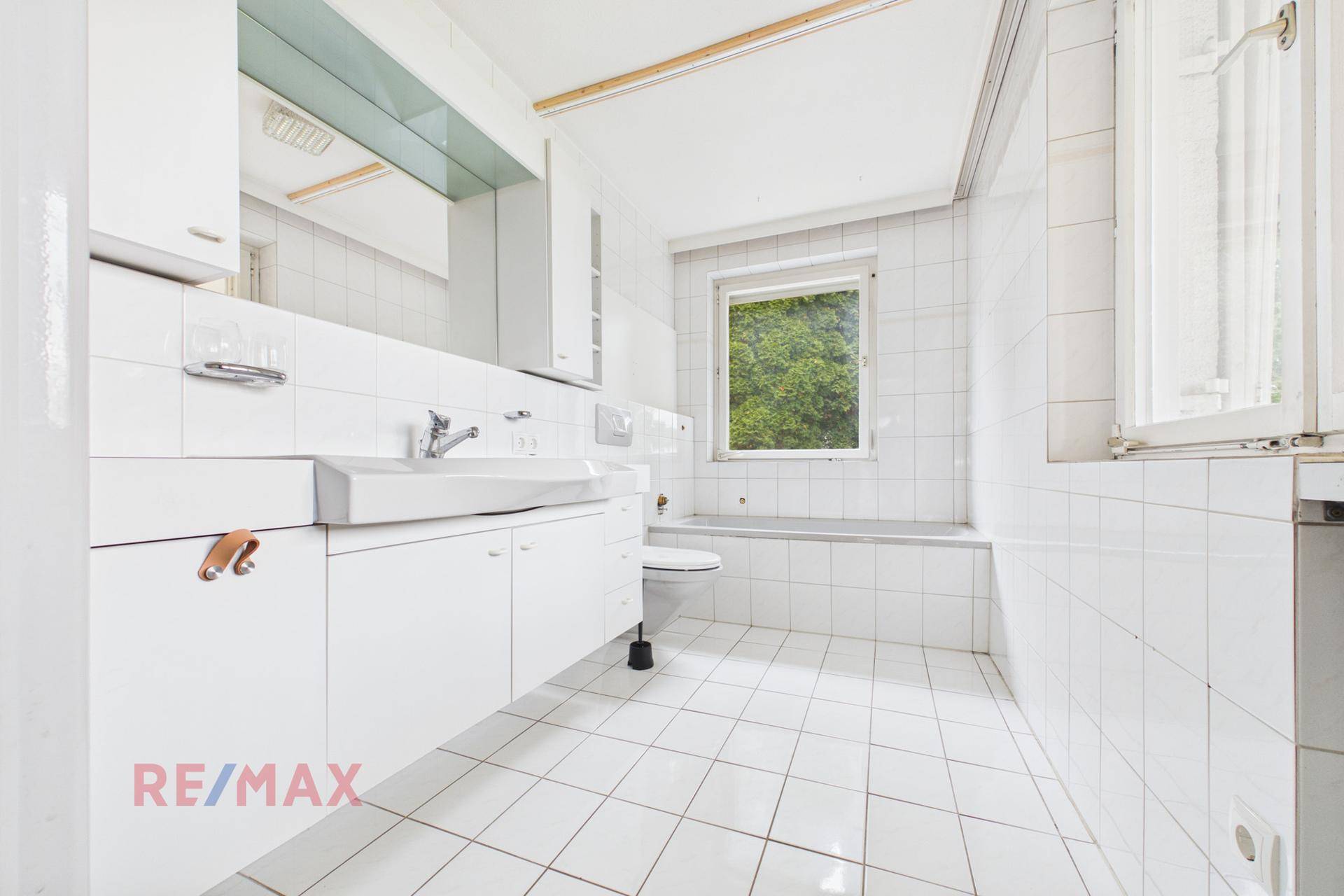 Helles Badezimmer mit weißen Fliesen, Badewanne und Fenster zum Garten.