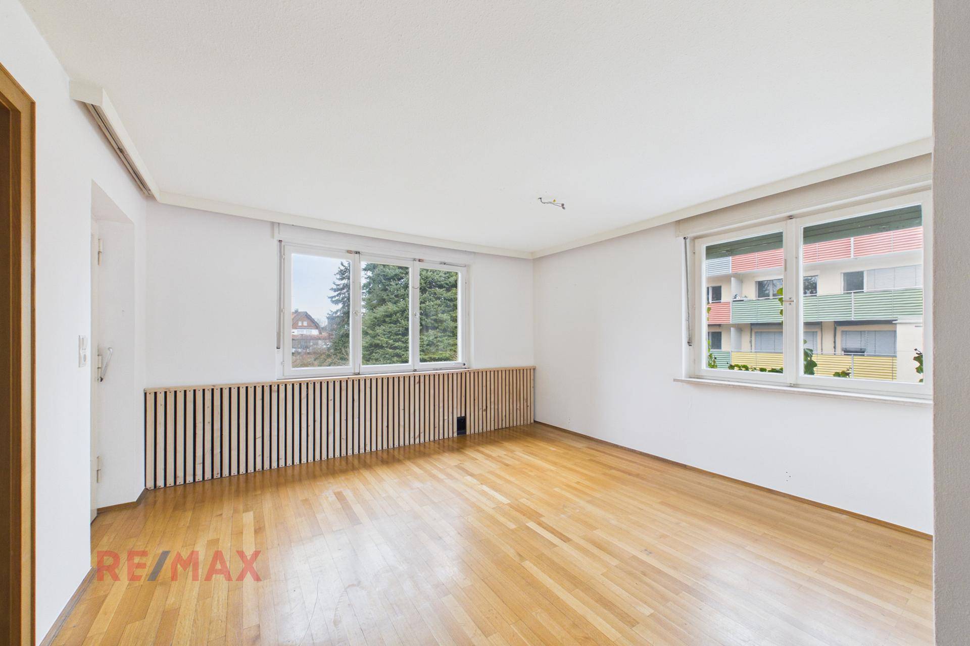 Helles Zimmer mit Parkettboden und großen Fenstern, die viel Tageslicht hereinlassen.