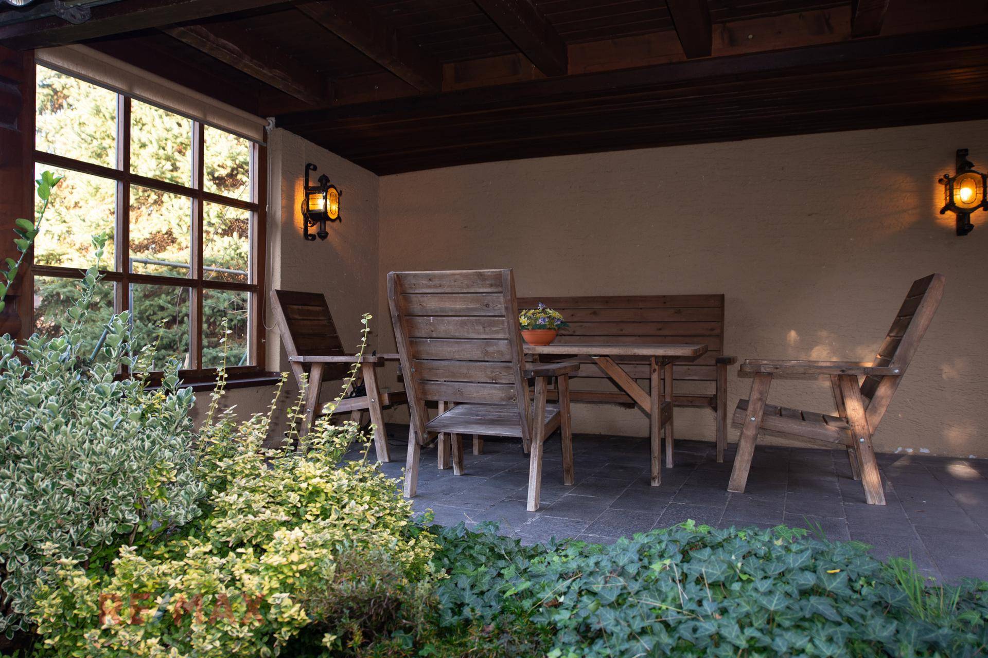 Überdachte Terrasse mit Holztisch und Stühlen, umgeben von Pflanzen.