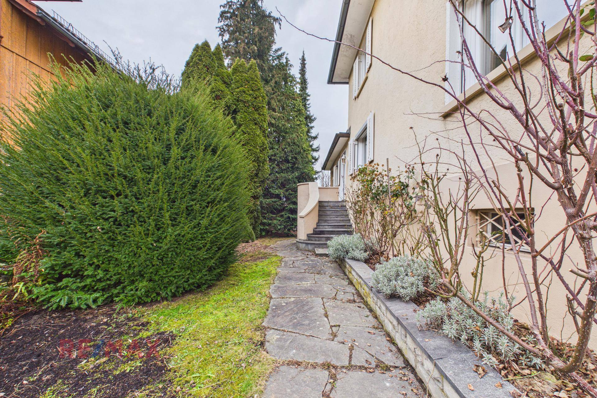 Steinplattenweg im Garten, umgeben von Sträuchern und Bäumen, führt zum Haus.