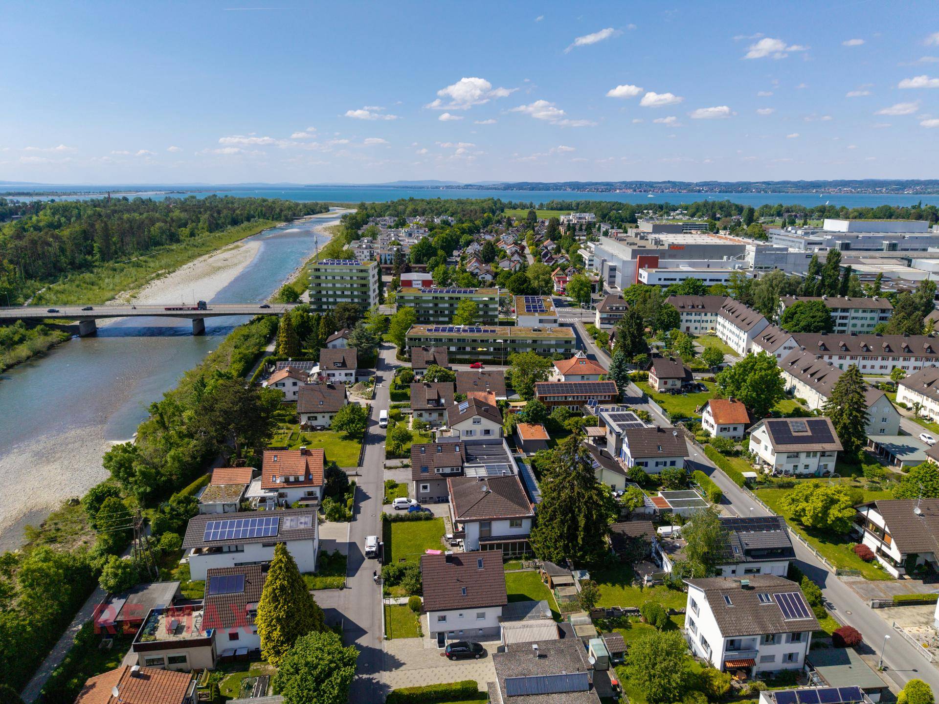 Weitläufige Luftaufnahme einer Wohnsiedlung mit Fluss, Brücke und See im Hintergrund unter blauem Himmel.