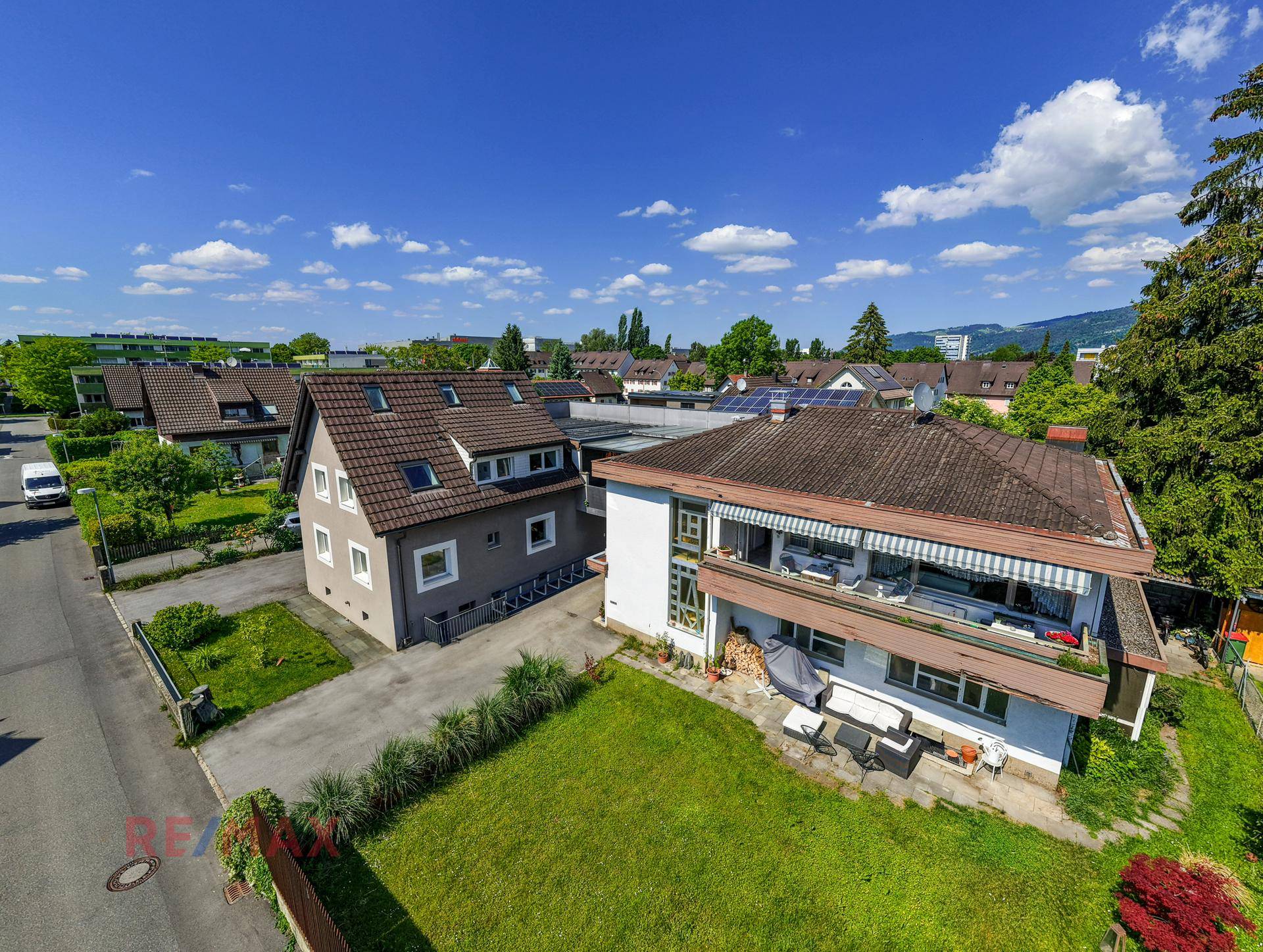 Panoramablick auf zwei Wohnhäuser mit Gärten und umliegender Wohnbebauung unter strahlend blauem Himmel.