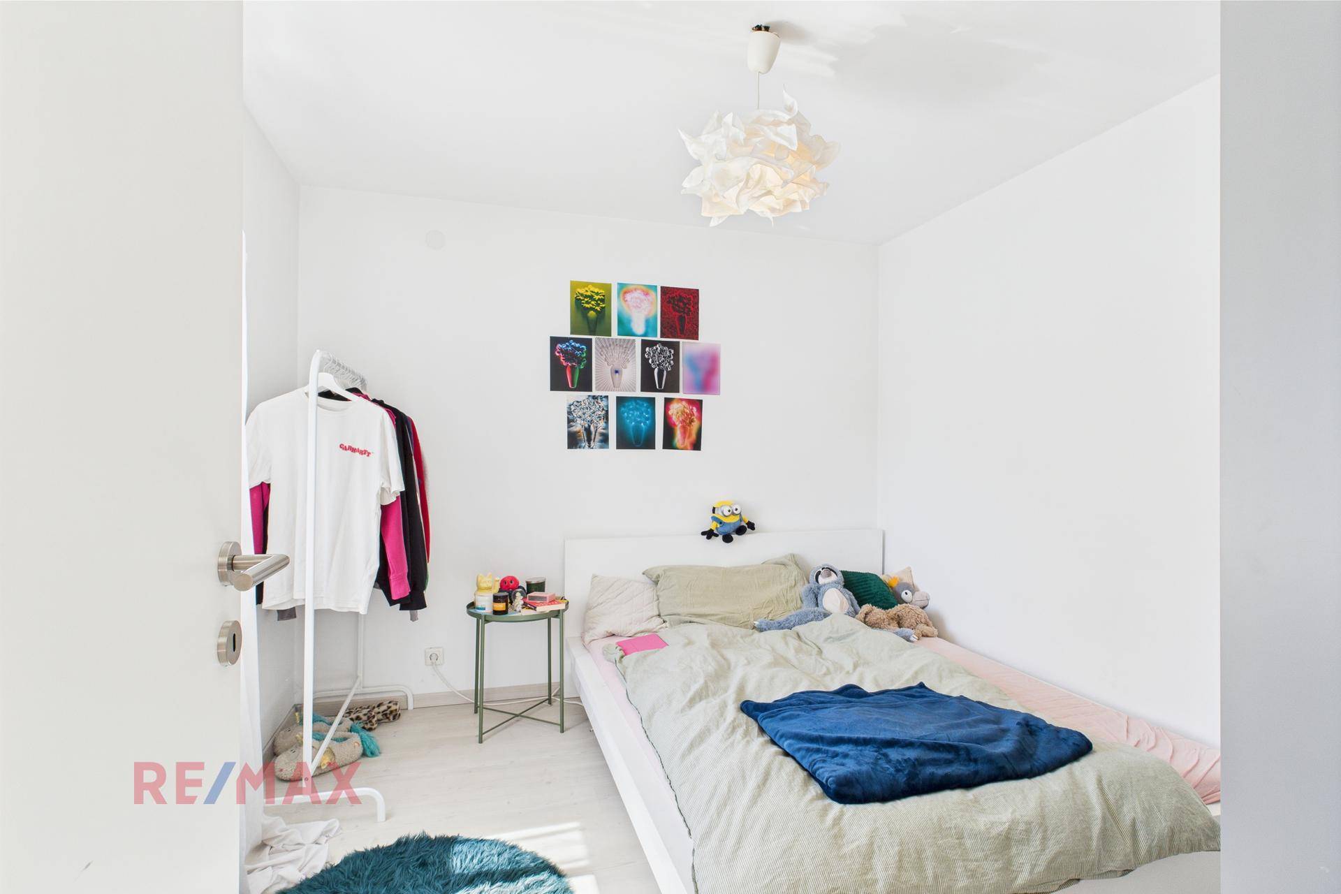 Schlichtes Schlafzimmer mit Einzelbett, weißem Kleiderständer und farbigen Bildern an der Wand.