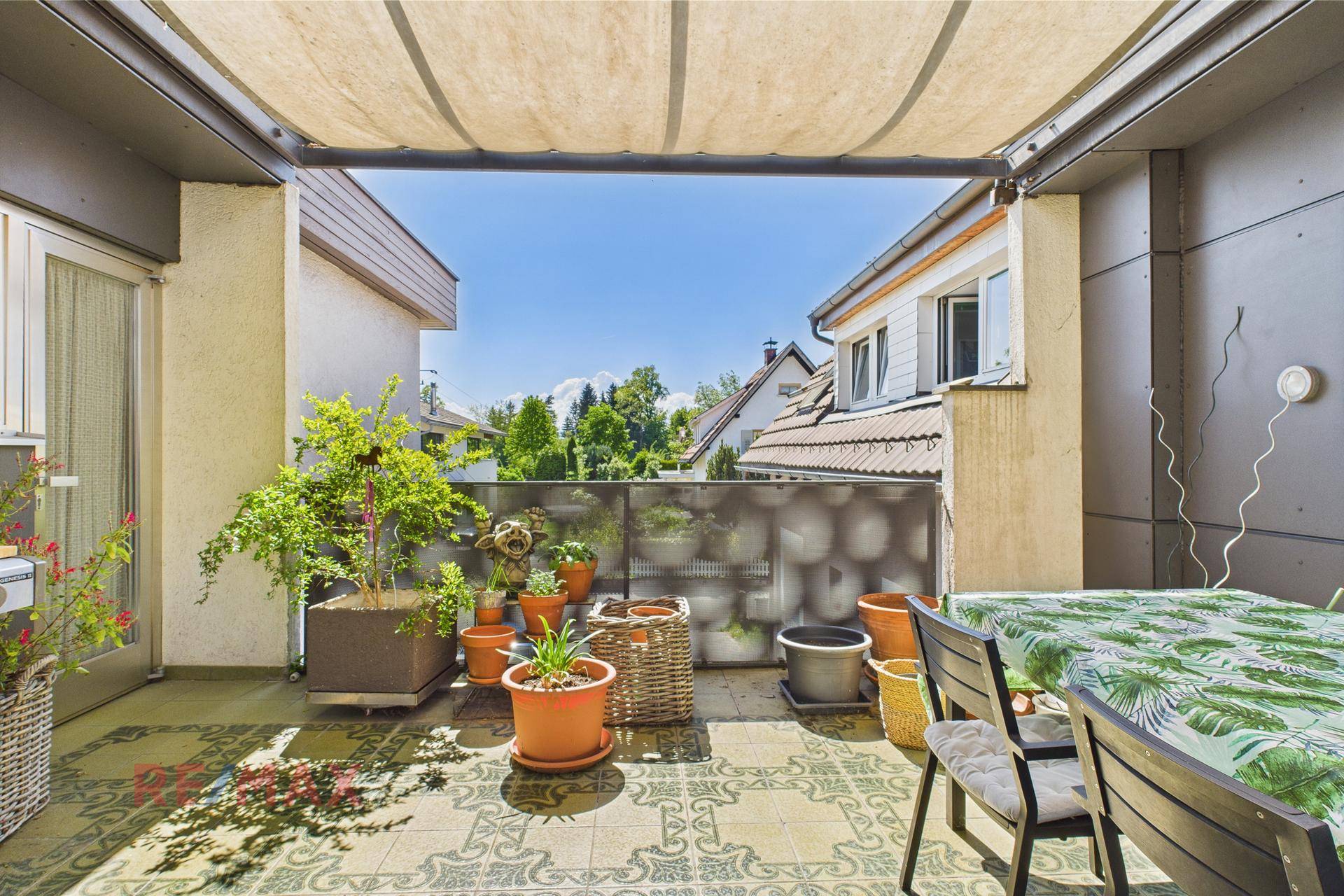 Überdachte Terrasse mit gemusterten Fliesen, Esstisch und vielen Topfpflanzen unter blauem Himmel.