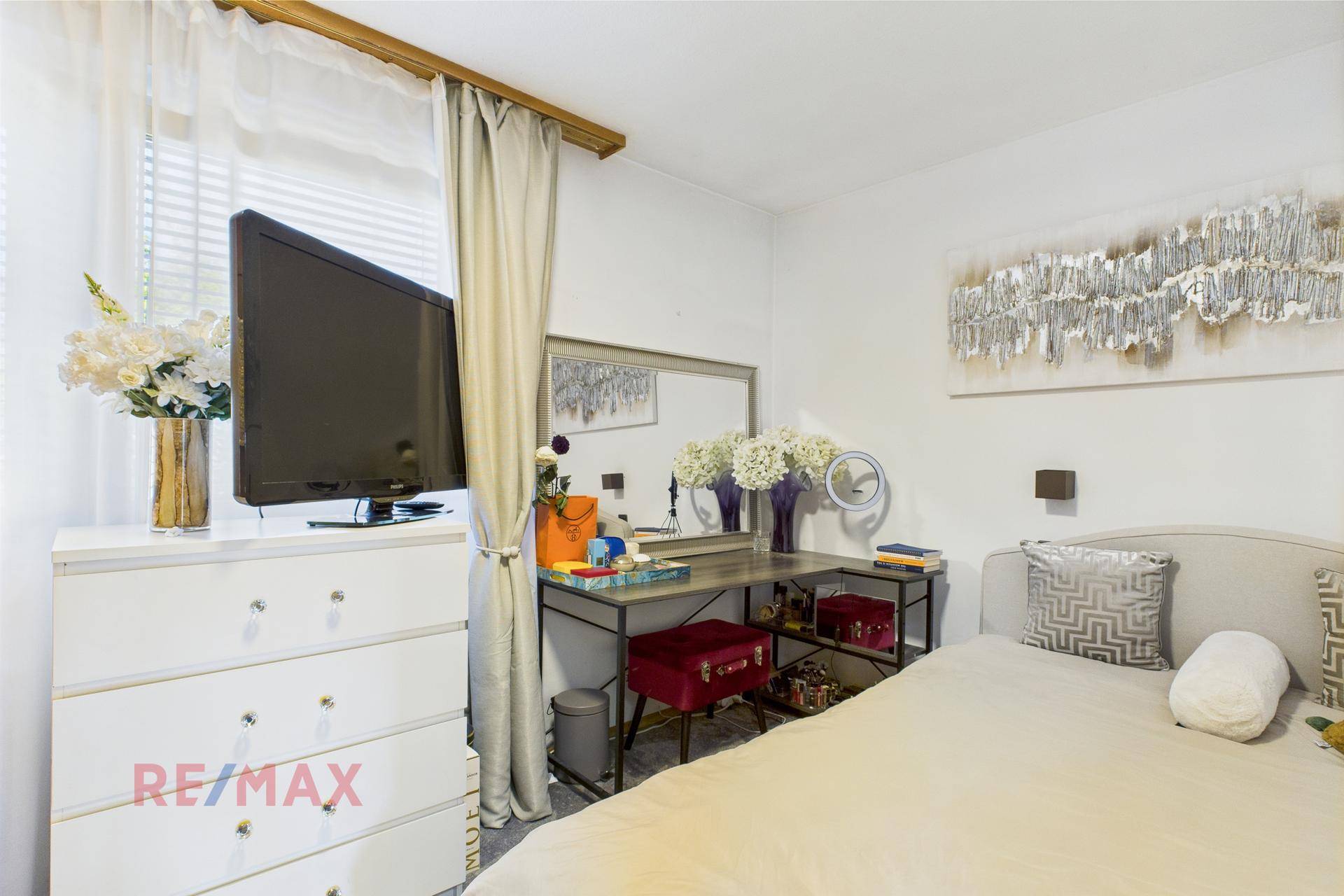 Schlafzimmer mit Bett, Kommode, Fernseher und Schminktisch vor einem großen Fenster.