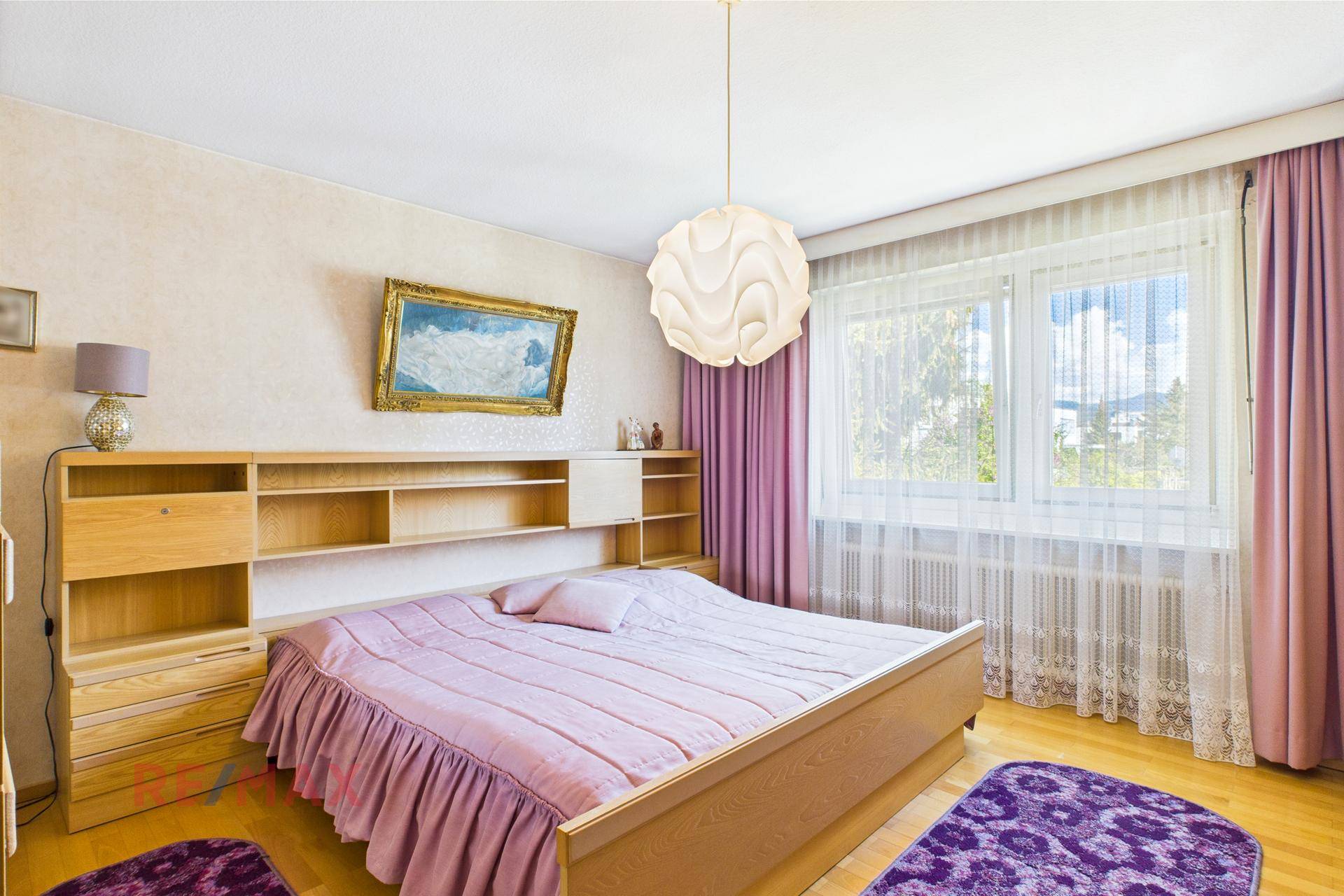 Geräumiges Schlafzimmer mit Doppelbett, Holzregal und großen Fenstern mit lila Vorhängen.
