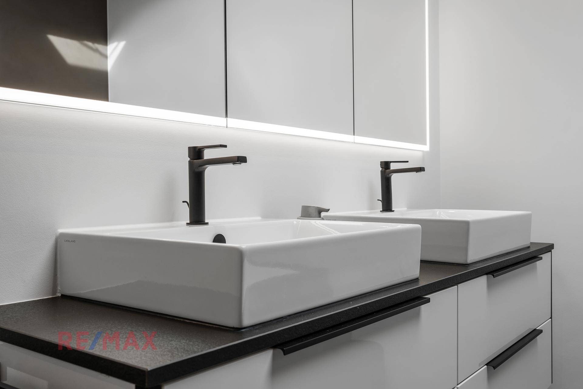 Modernes Badezimmer mit Doppelwaschbecken, schwarzen Armaturen und beleuchtetem Spiegelschrank.