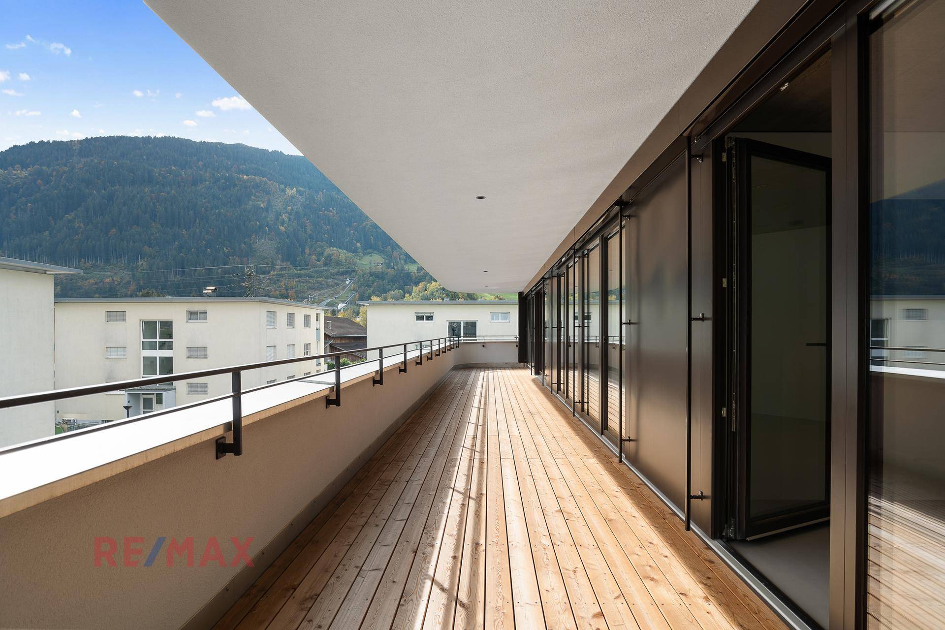 Langer Holzbalkon mit Blick auf die umliegende Berglandschaft und benachbarte Häuser.