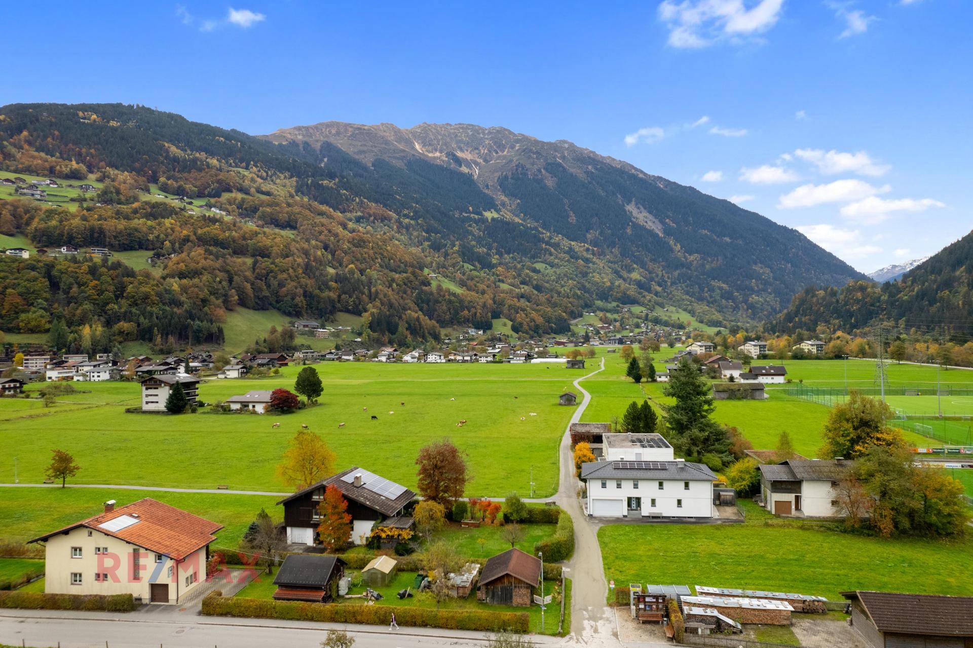 Weitläufige Landschaft mit grünen Wiesen, Häusern und majestätischen Bergen im Hintergrund.