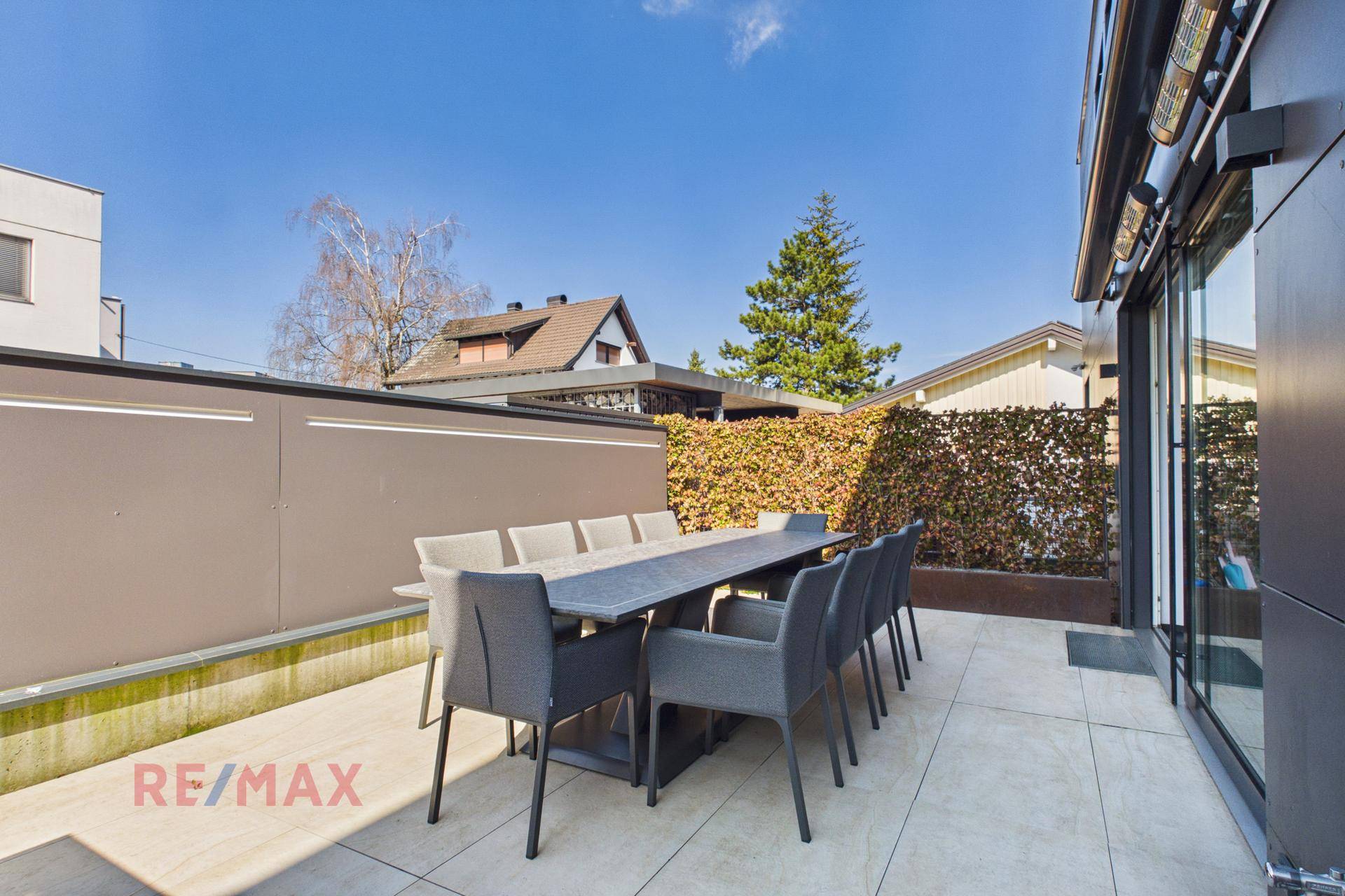 Sonniger Terrassenbereich mit großem Esstisch und Stühlen, umgeben von einer grünen Hecke.