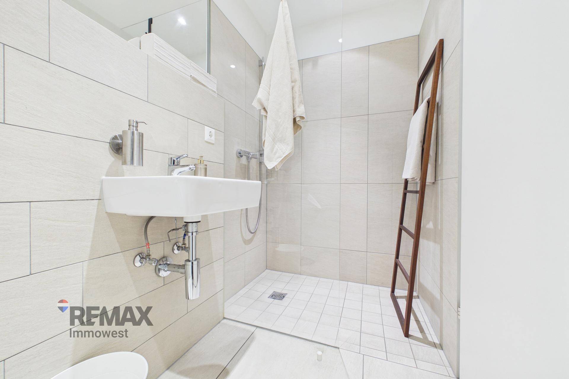 Modernes Badezimmer mit begehbarer Dusche, wandmontiertem Waschbecken und hellen Fliesen.