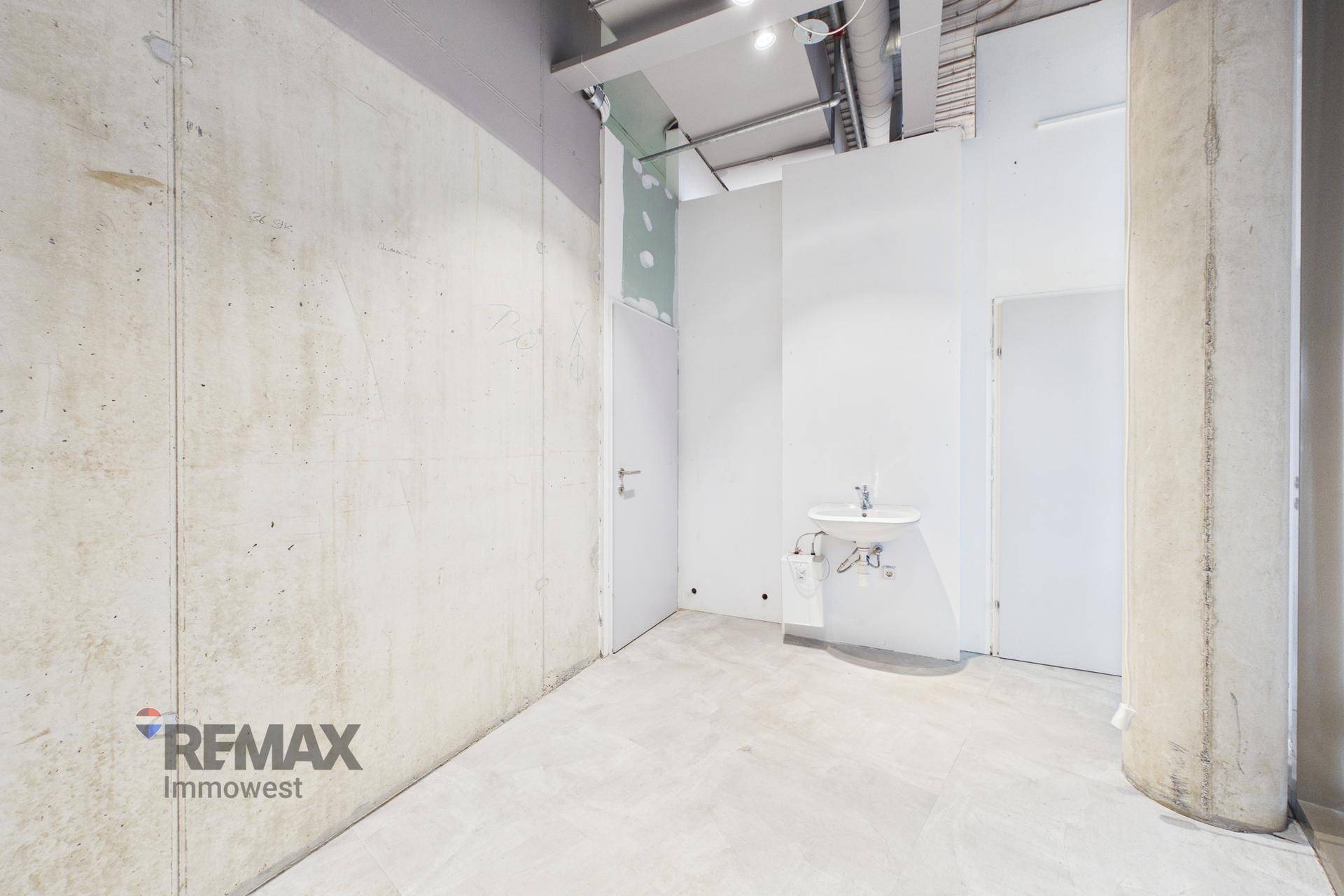 Kleiner Raum mit Waschbecken, Betonwand und sichtbaren Deckeninstallationen, vorbereitet für eine Küchenzeile oder Nasszelle.
