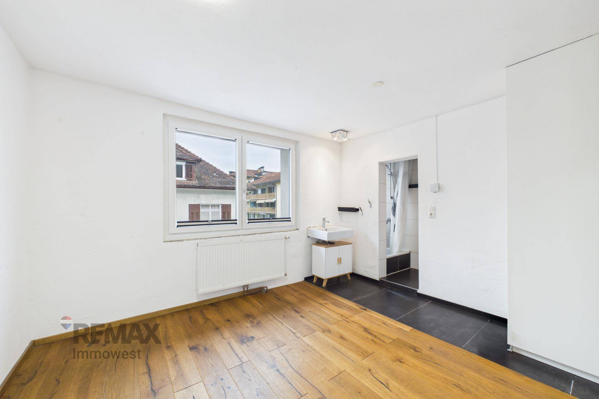 Zimmer mit Holzboden, integriertem Waschbecken und einem Duschbereich, beleuchtet durch ein Fenster.