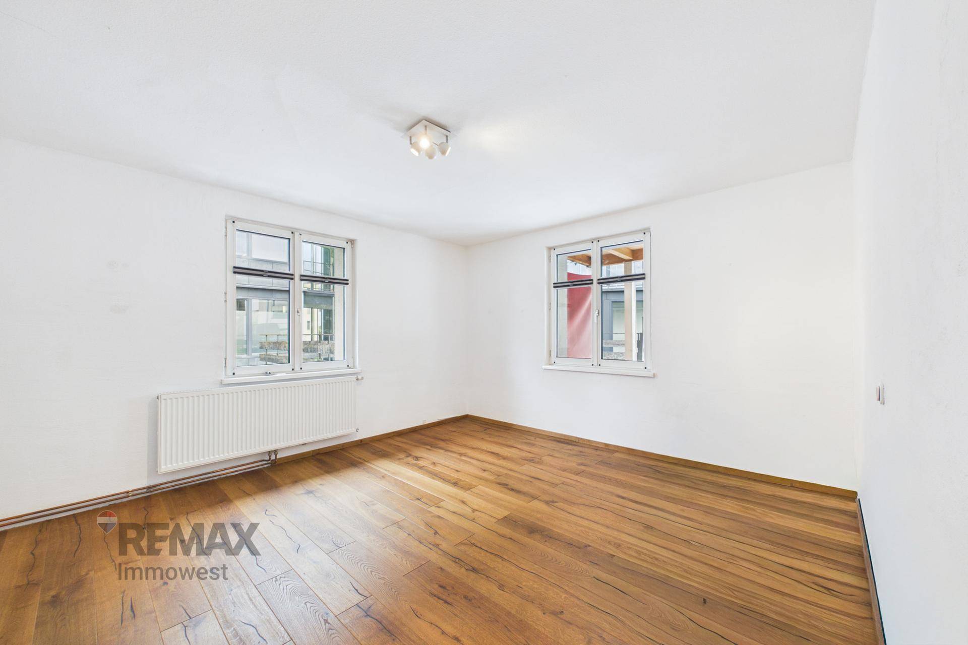 Leeres Zimmer mit Holzboden, weißen Wänden und zwei großen Fenstern für viel Tageslicht.