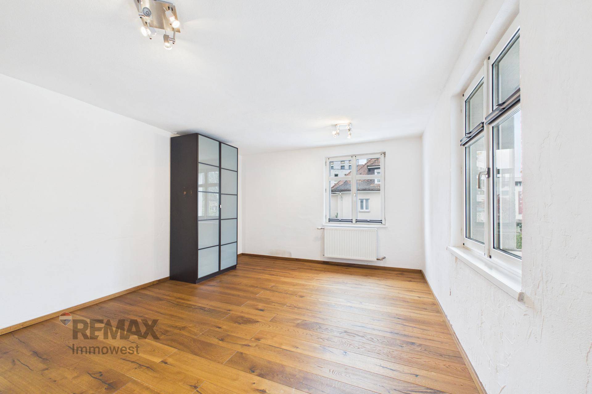 Leeres Zimmer mit Holzboden, weißen Wänden und einem Fenster, das Tageslicht hereinlässt.