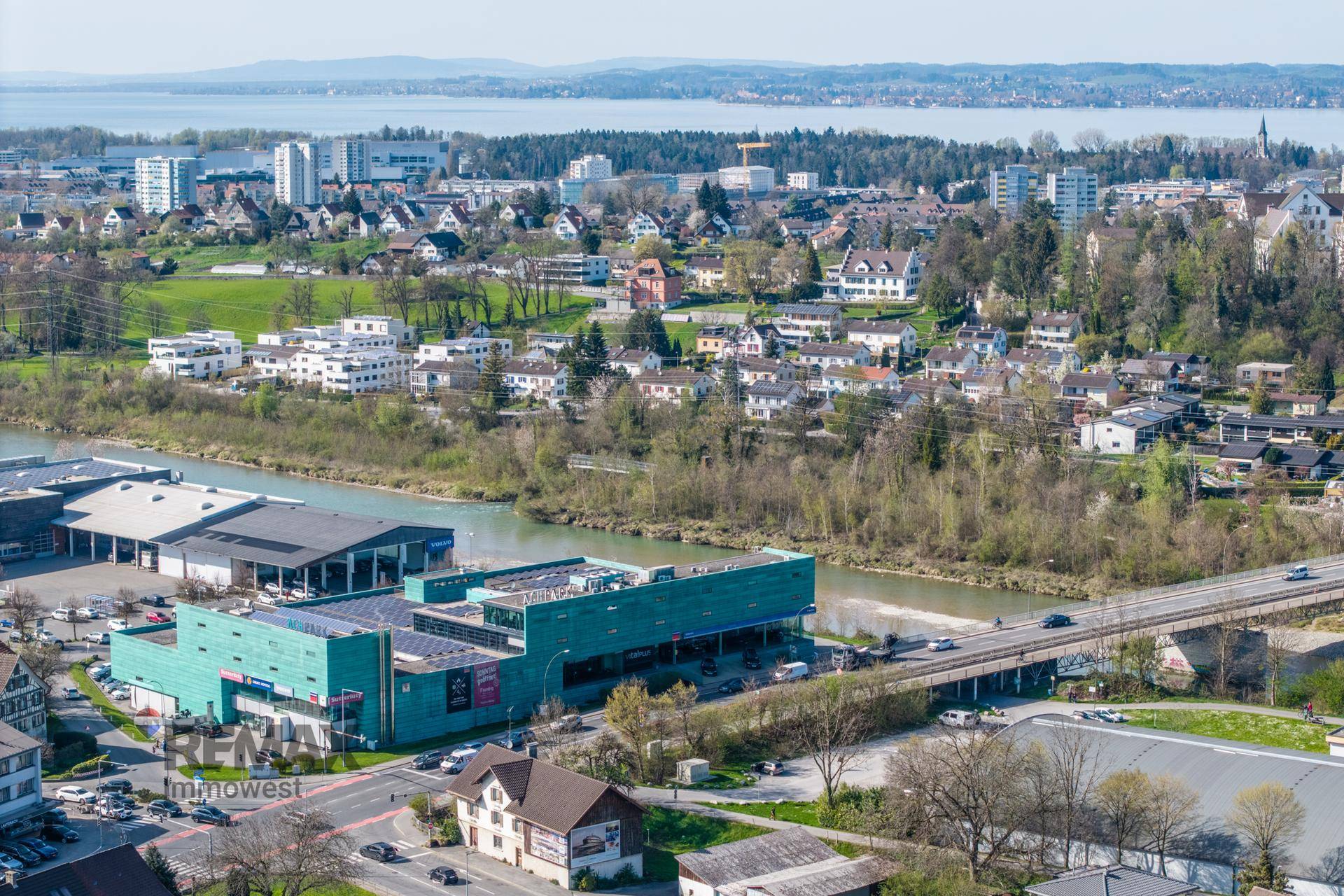 Weitläufige Luftaufnahme des Gewerbegebiets mit Fluss, See und Wohnsiedlungen im Hintergrund unter blauem Himmel.