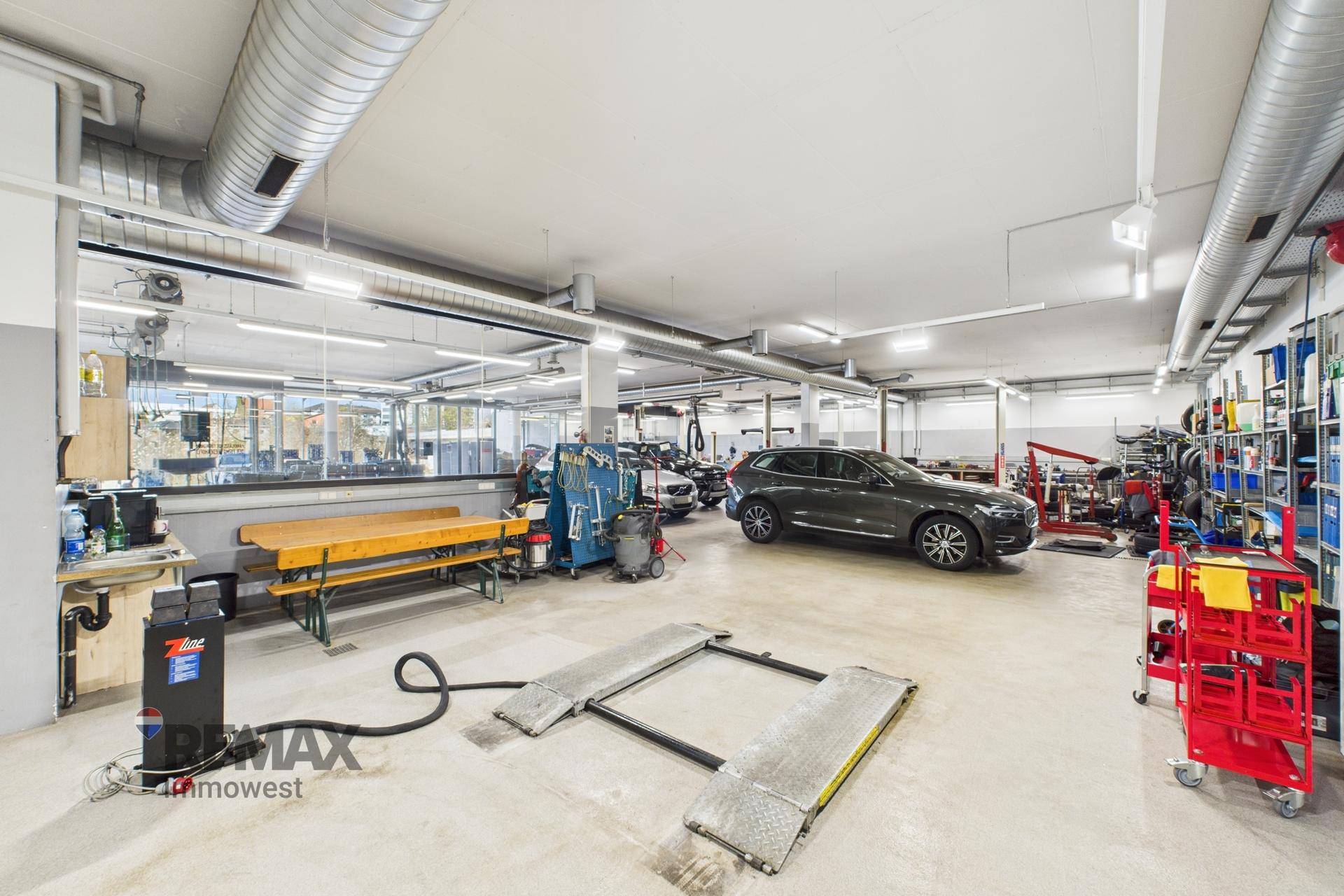 Werkstatt mit Fahrzeug auf einer Hebebühne, Werkbänken und Lüftungsrohren an der Decke.