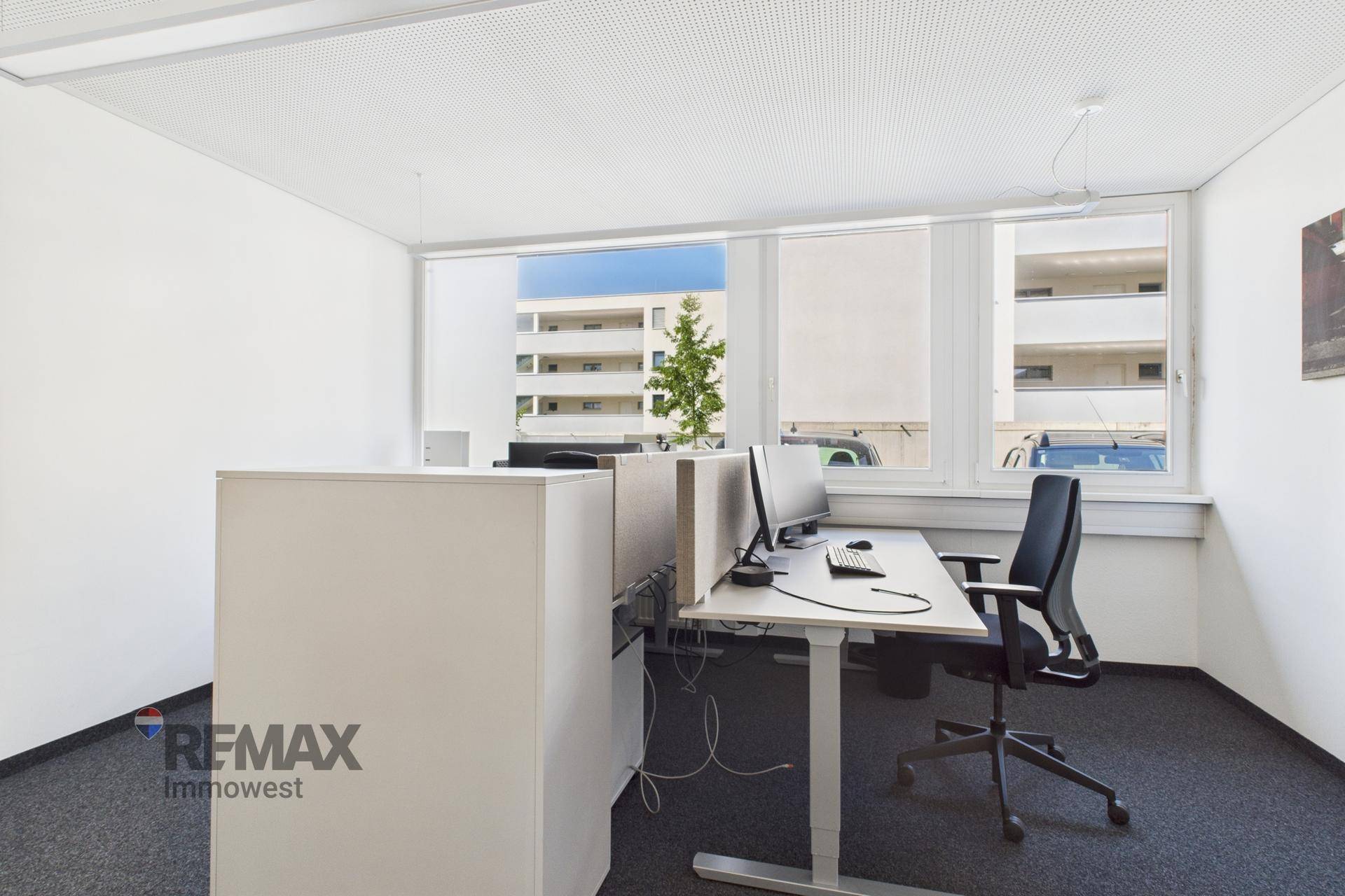 Moderner Arbeitsplatz in einem hellen Büro mit großen Fenstern und Blick auf umliegende Gebäude.