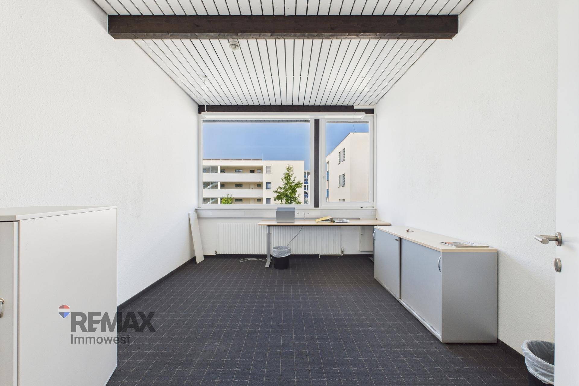 Geräumiges, leeres Büro mit großen Fenstern, die viel Licht hereinlassen, und einem einfachen Schreibtisch.