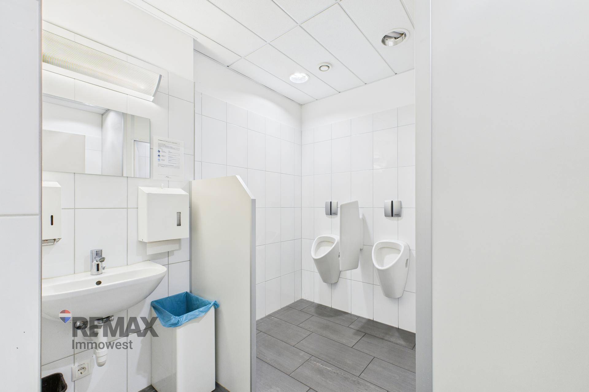 Herren-WC mit zwei Urinalen, Waschbecken und weißen Wandfliesen.