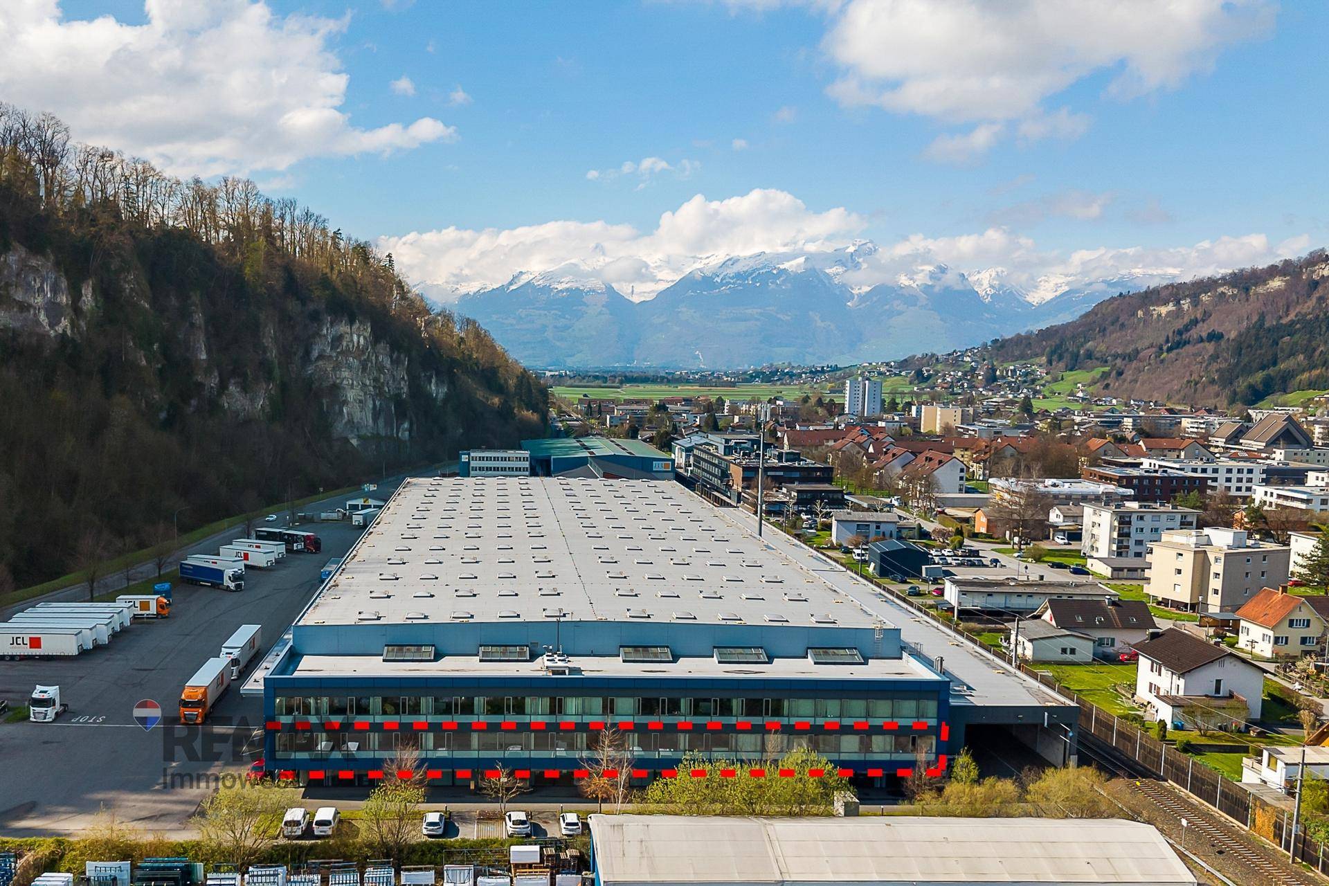 Weitläufige Luftaufnahme eines Industriegebiets mit großem Gebäude und Alpenpanorama.