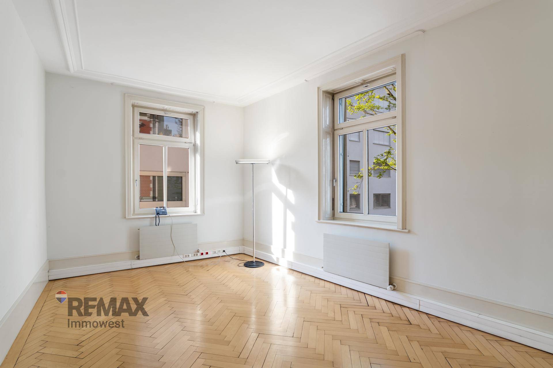 Leeres Büro mit Fischgrätparkett, hohen Decken und großen Fenstern, die viel Tageslicht hereinlassen.