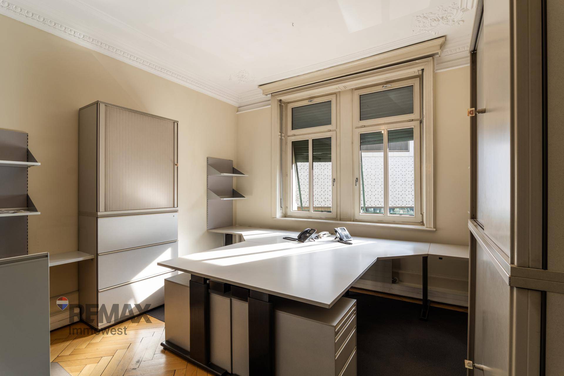 Arbeitsbereich mit Schreibtisch, Büromöbeln und großen Fenstern, die viel Tageslicht hereinlassen.
