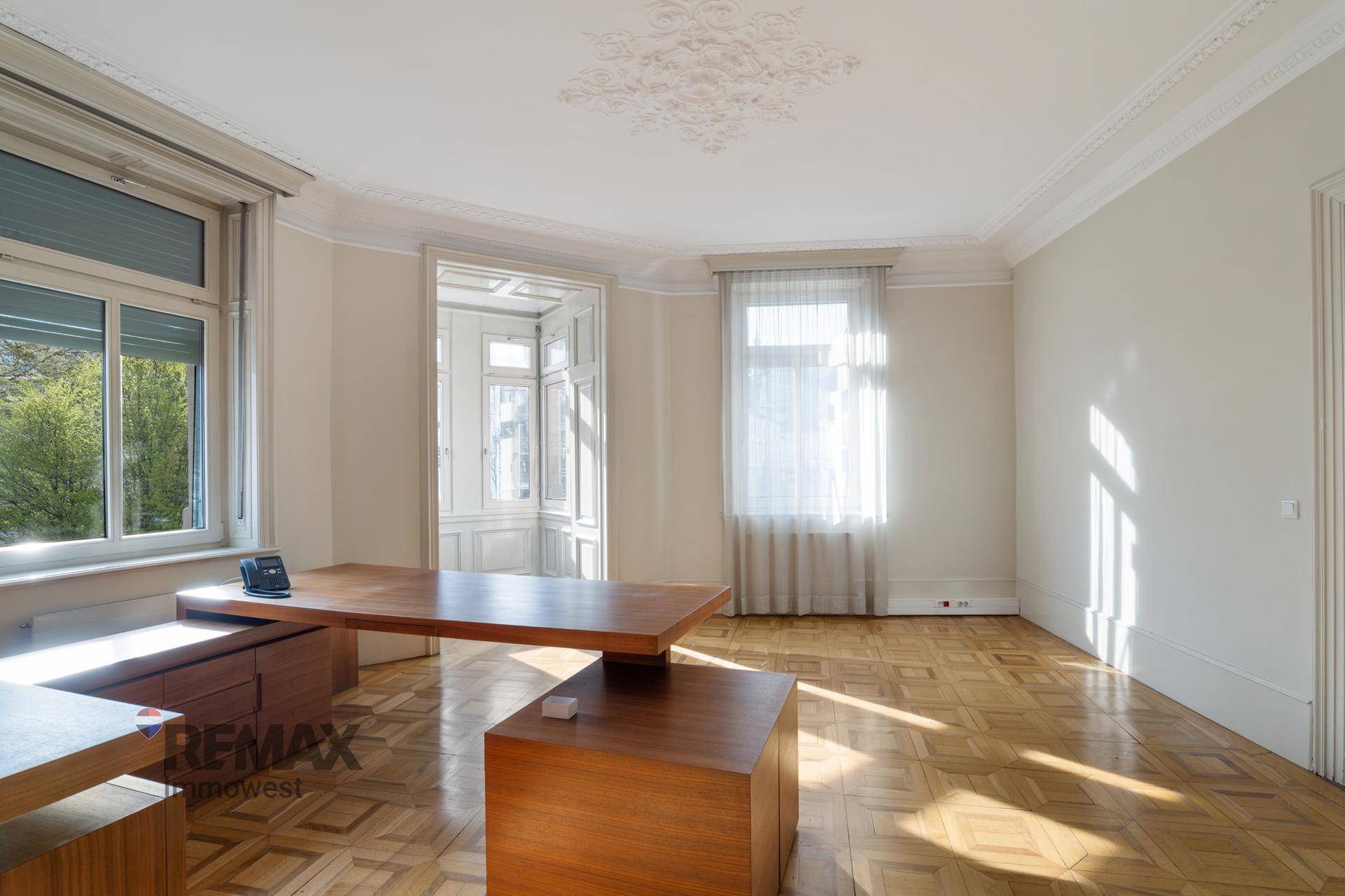 Geräumiges Büro mit Fischgrätparkett, hohen Decken und einem großen Schreibtisch aus Holz.