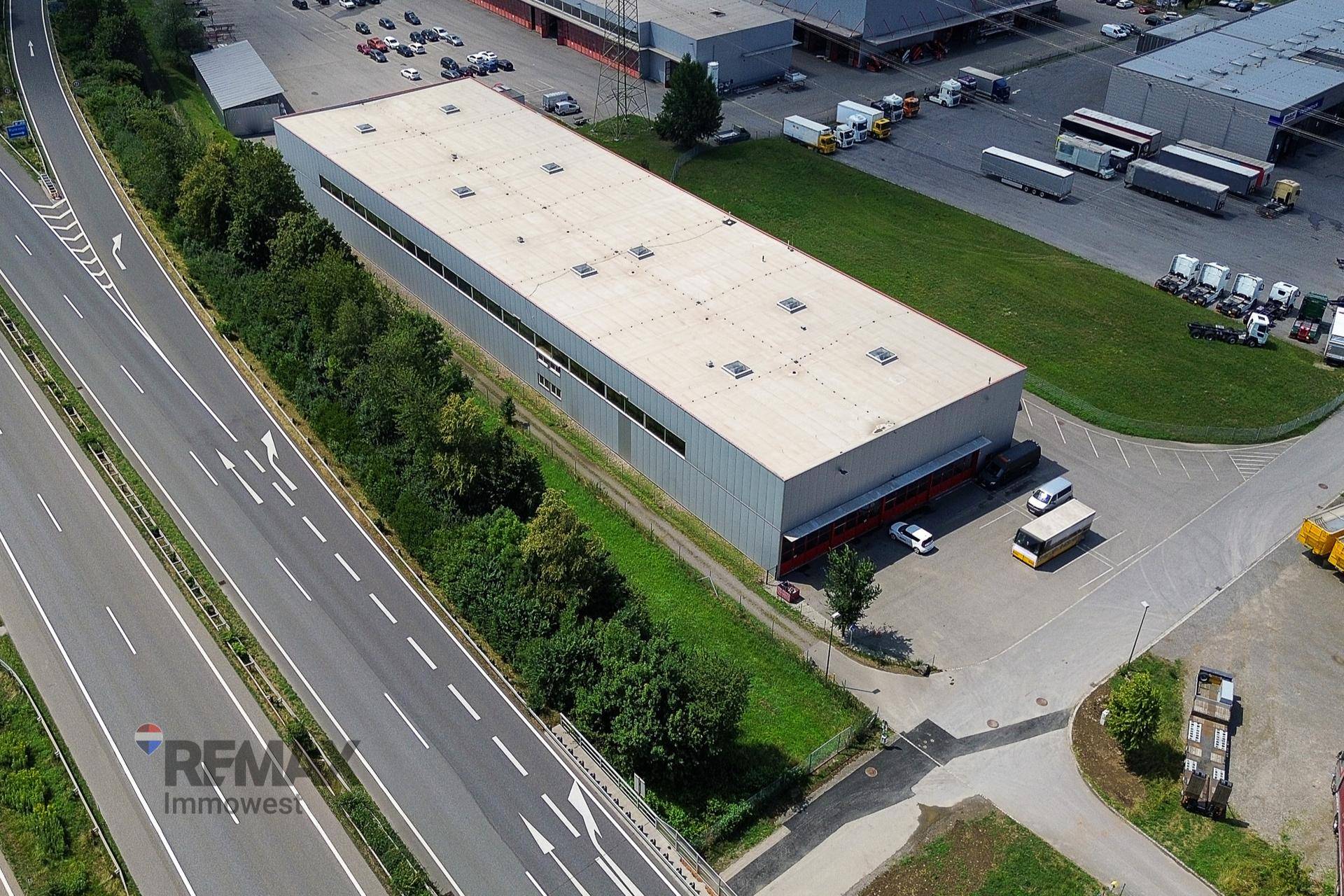 Luftaufnahme eines großen Industriegebäudes neben einer Autobahn, umgeben von Grünflächen und Parkplätzen.