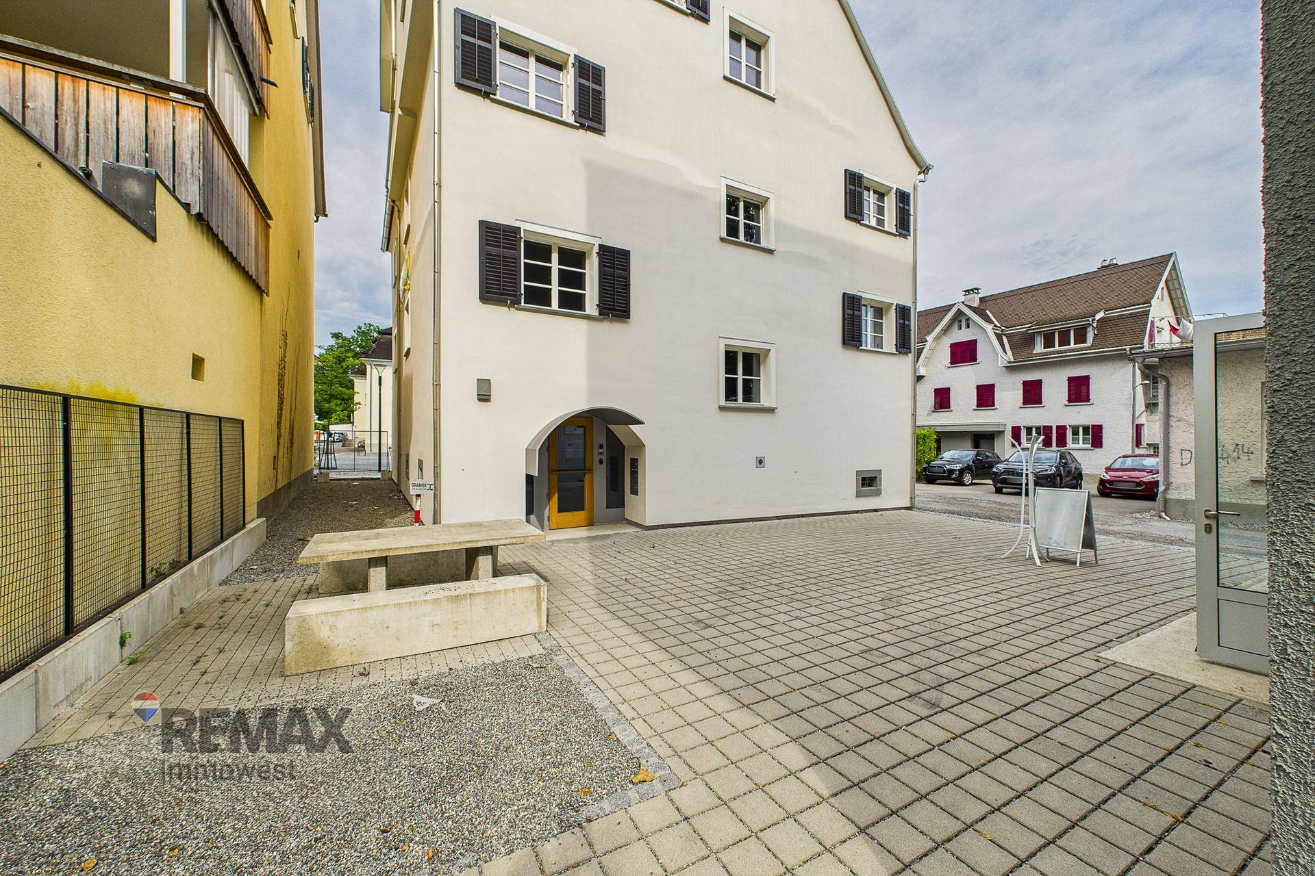 Außenansicht des Gebäudes mit gepflastertem Vorplatz und Sitzgelegenheiten im Freien.