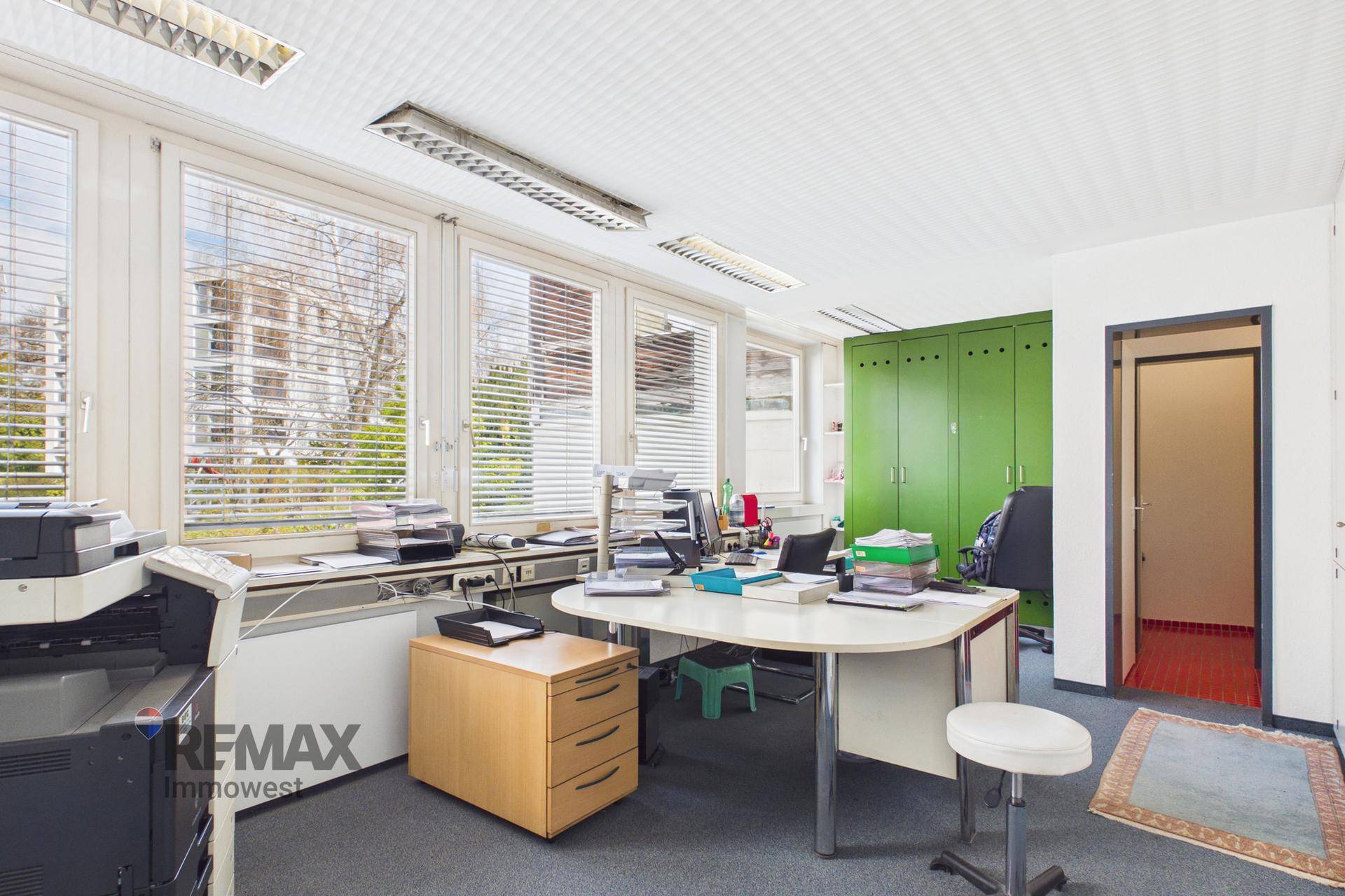 Arbeitsplatz in einem Büro mit Schreibtisch, Drucker, grünen Schränken und Fenstern mit Jalousien.